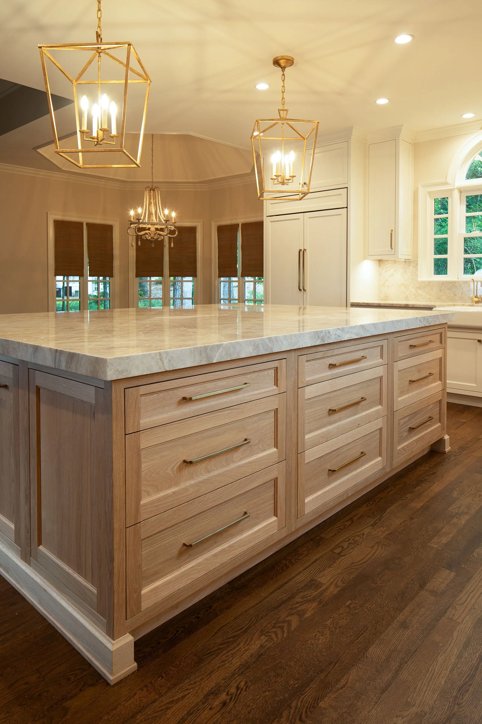 A kitchen island with a marble countertop, wooden drawers with gold handles, and hardwood flooring. In the background, there are windows with brown blinds, white cabinets, and various hanging light fixtures.