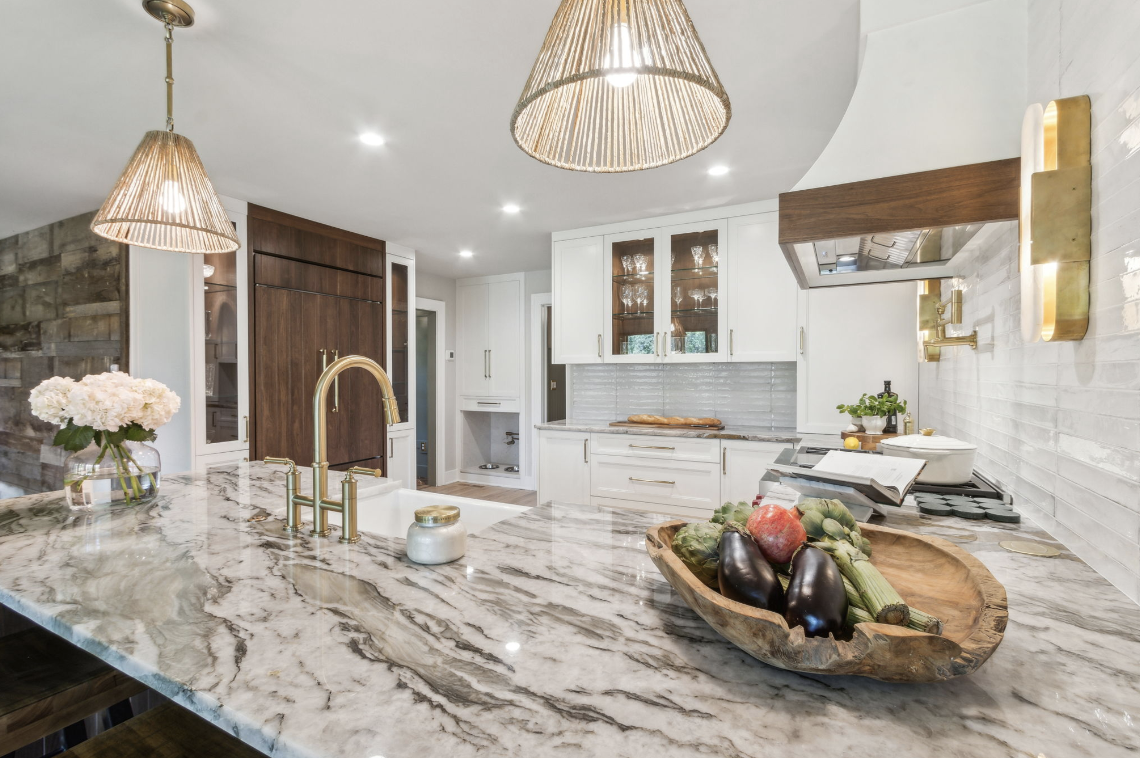 Modern kitchen with marble island, white cabinets, wooden accents, pendant lighting, and fresh vegetables in a wooden bowl on the counter.