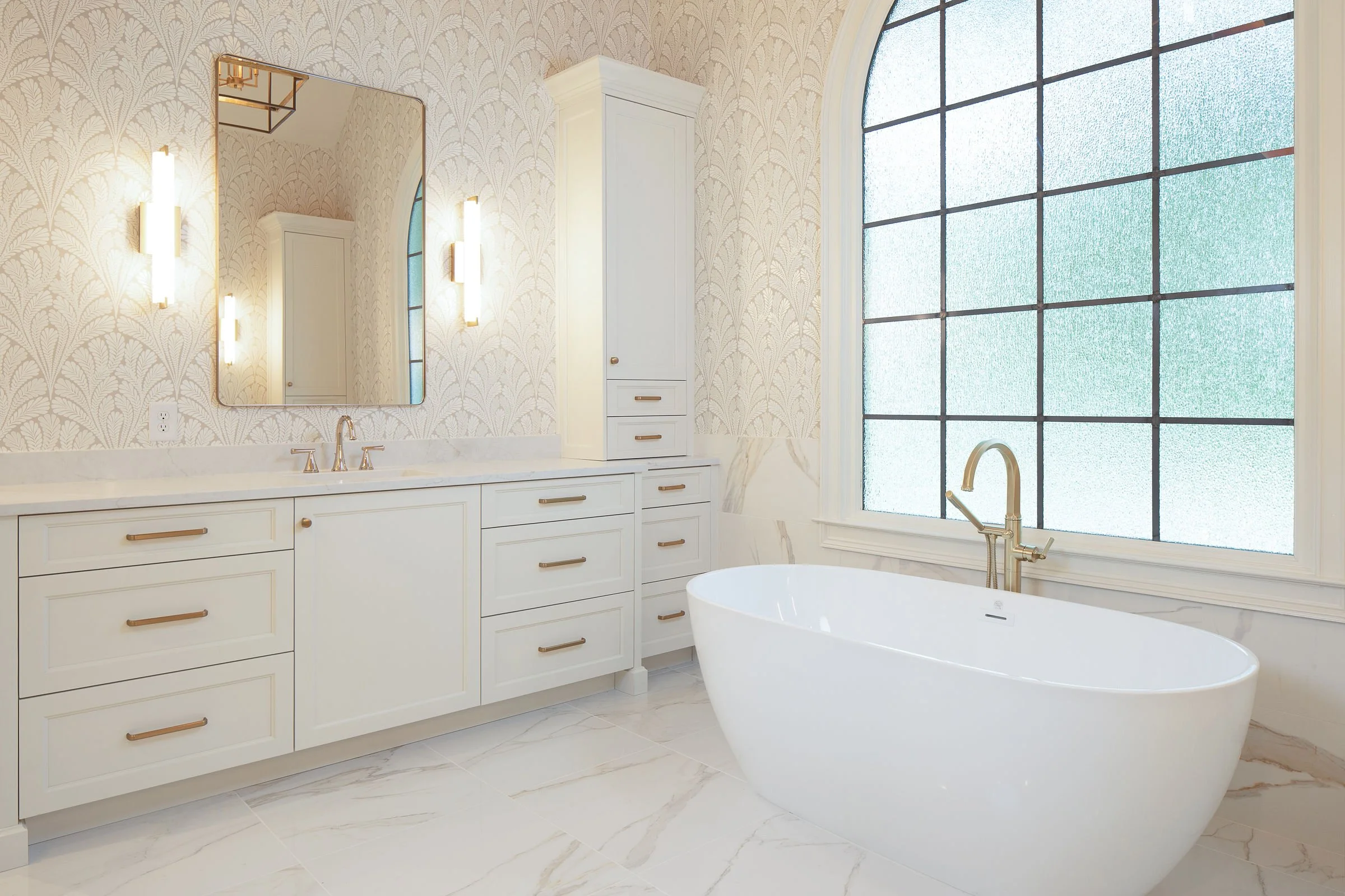 A bathroom with a white bathtub, gold fixtures, a large window, white cabinetry with gold handles, and a mirror on the wall.