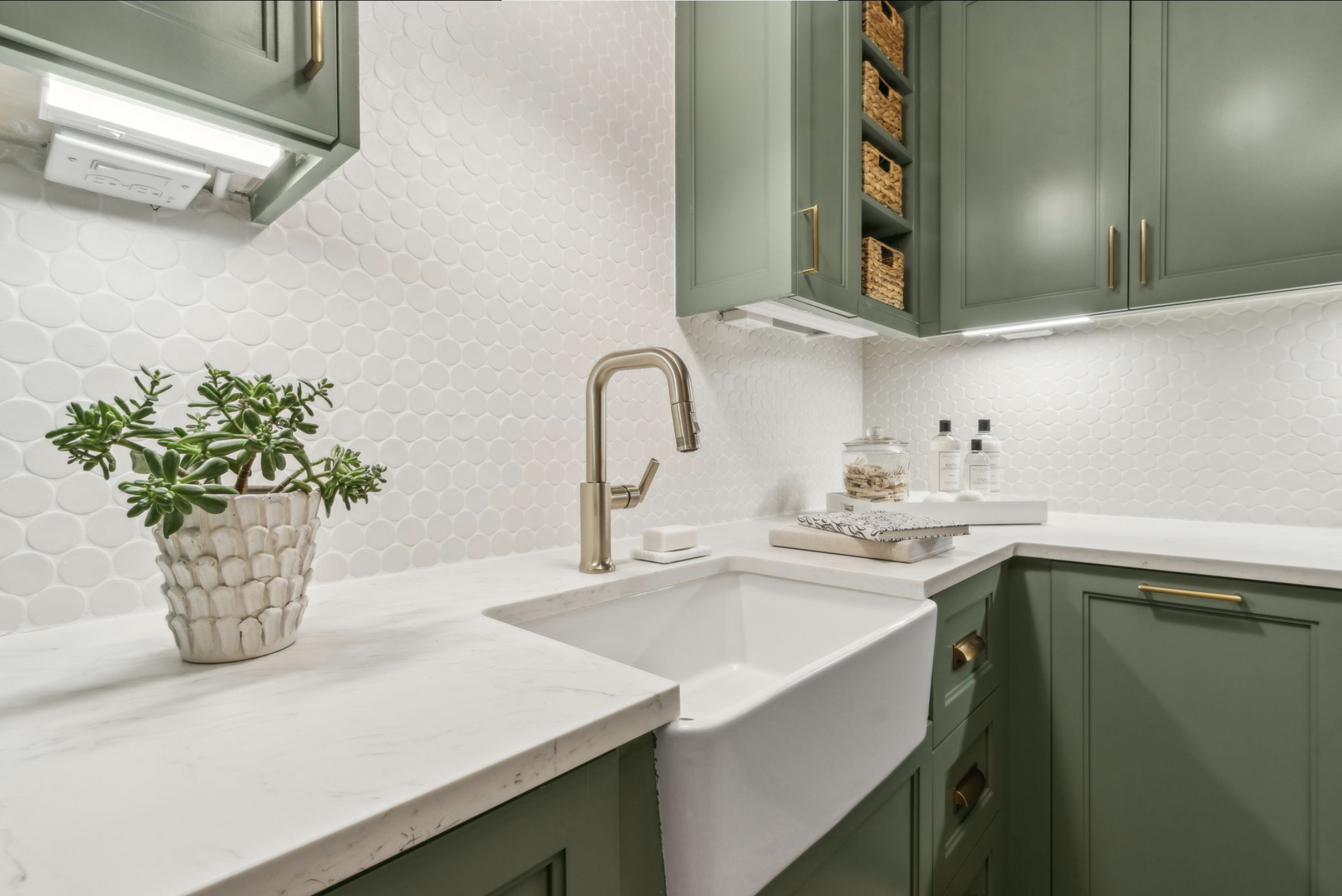 Clean kitchen sink with white subway tile backsplash, green cabinets with gold hardware, potted plant, and decorative items on the countertop.
