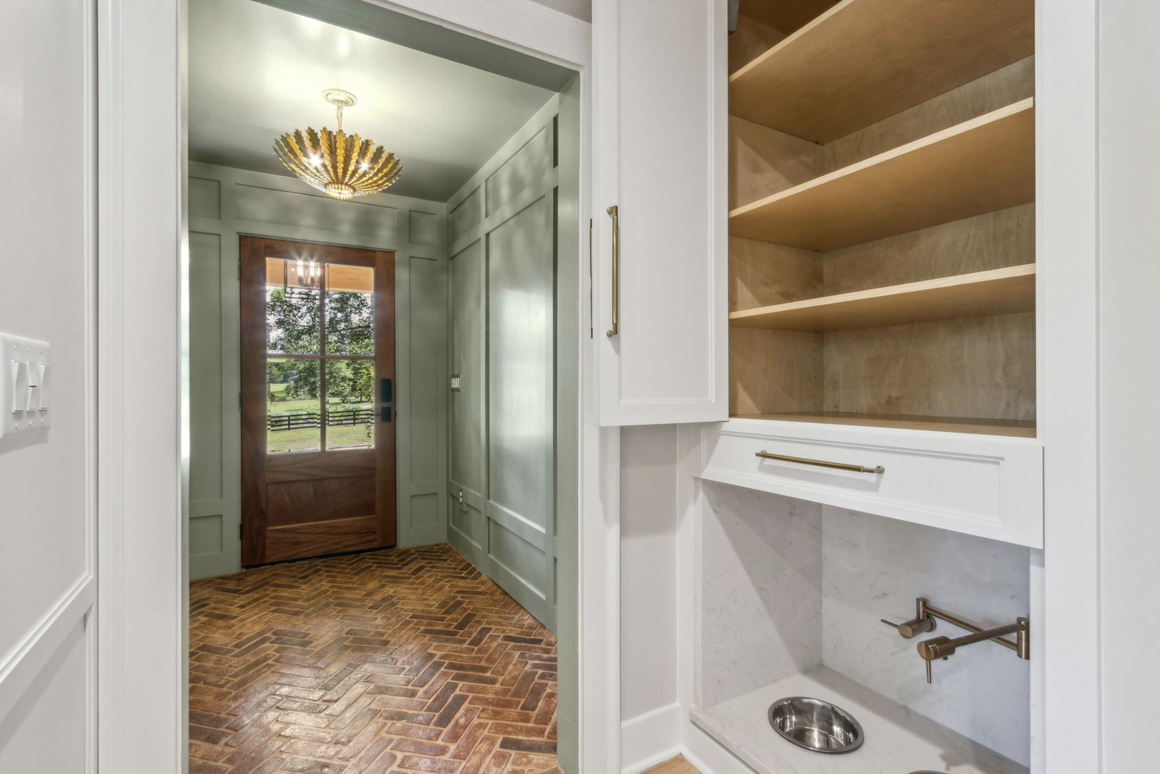 Entryway with a wooden door and a view of a green outdoor landscape. The floor is brick herringbone pattern. There is a chandelier hanging from the ceiling. On the right, built-in white cabinetry with shelves and a pet water bowl.