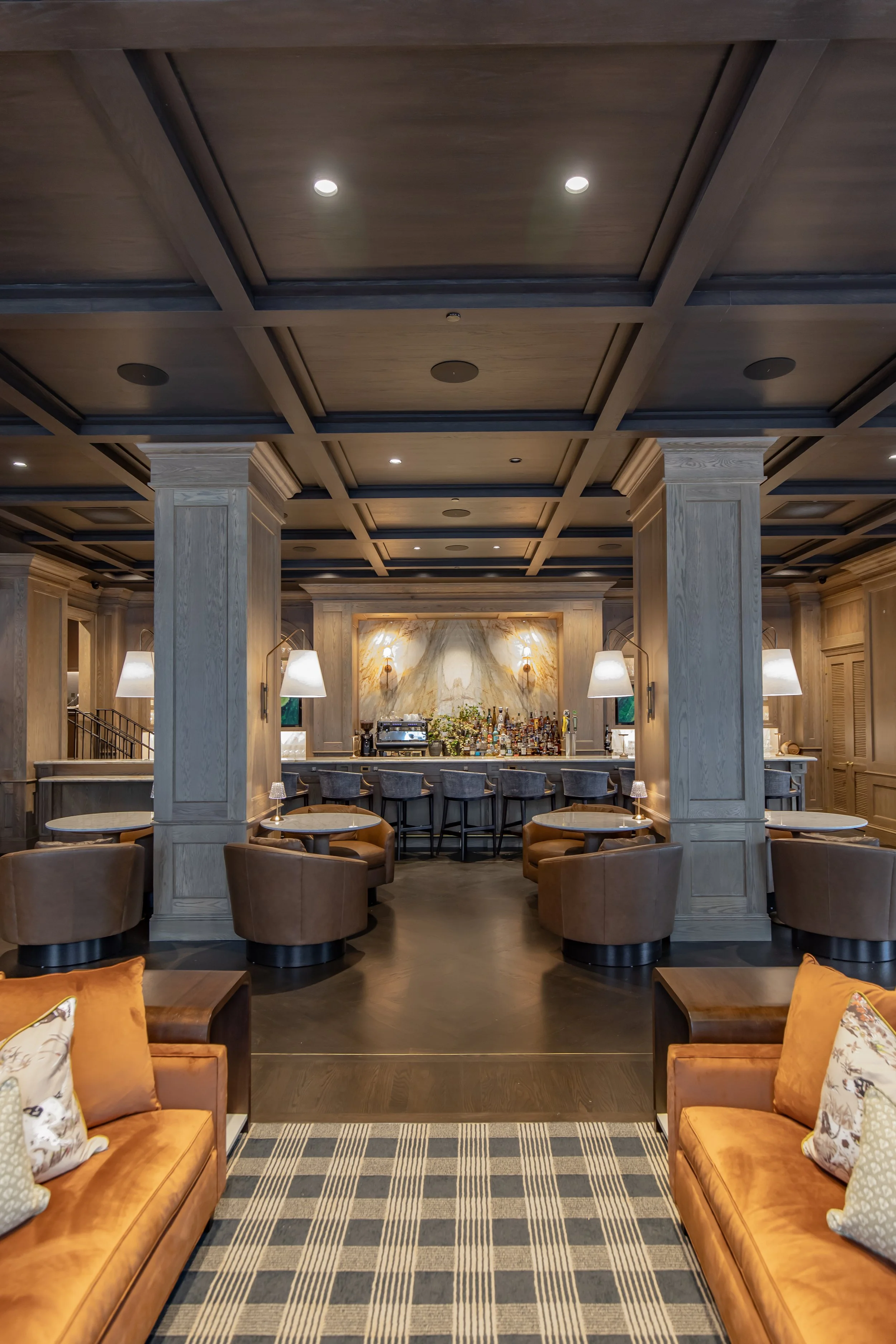 A lounge area with brown leather sofas and patterned pillows in the foreground, and a bar with a marble backsplash and barstools in the background, surrounded by wood-paneled walls and ceiling with recessed lighting.