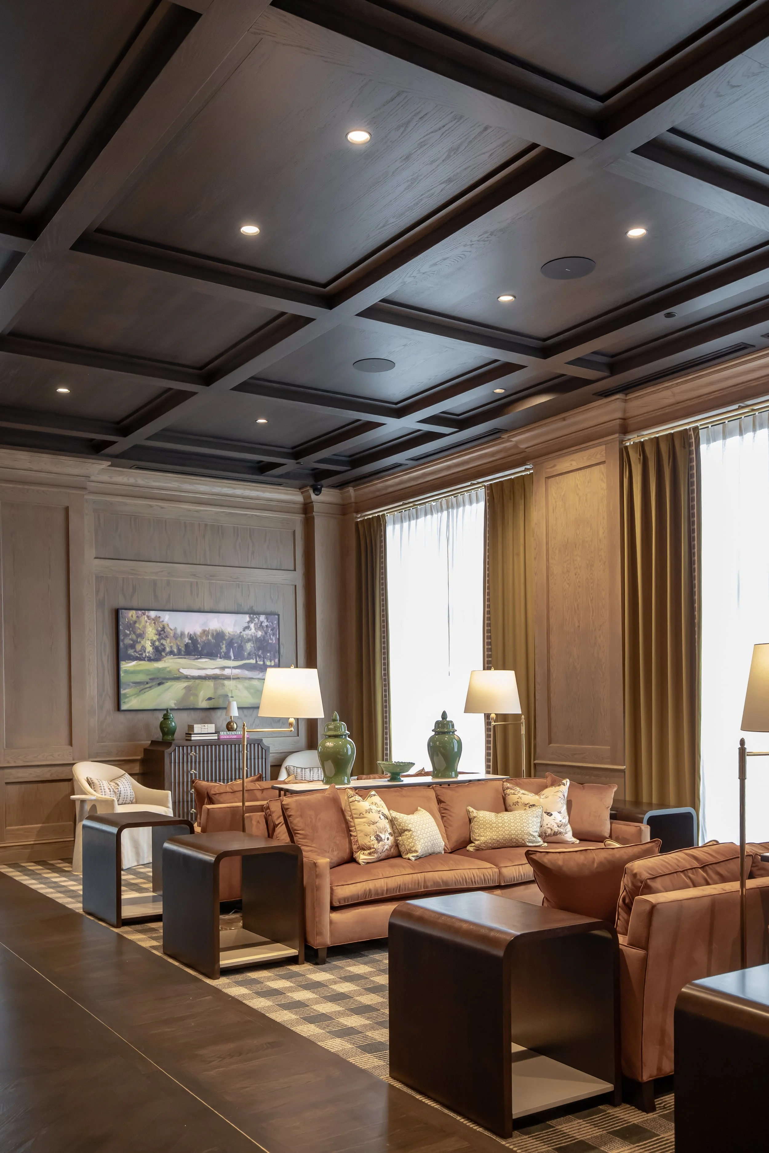 A luxurious living room with a coffered dark wood ceiling, beige curtains, a pink velvet sofa with patterned and solid throw pillows, green decorative vases, table lamps, and a framed landscape painting on light wood-paneled walls.
