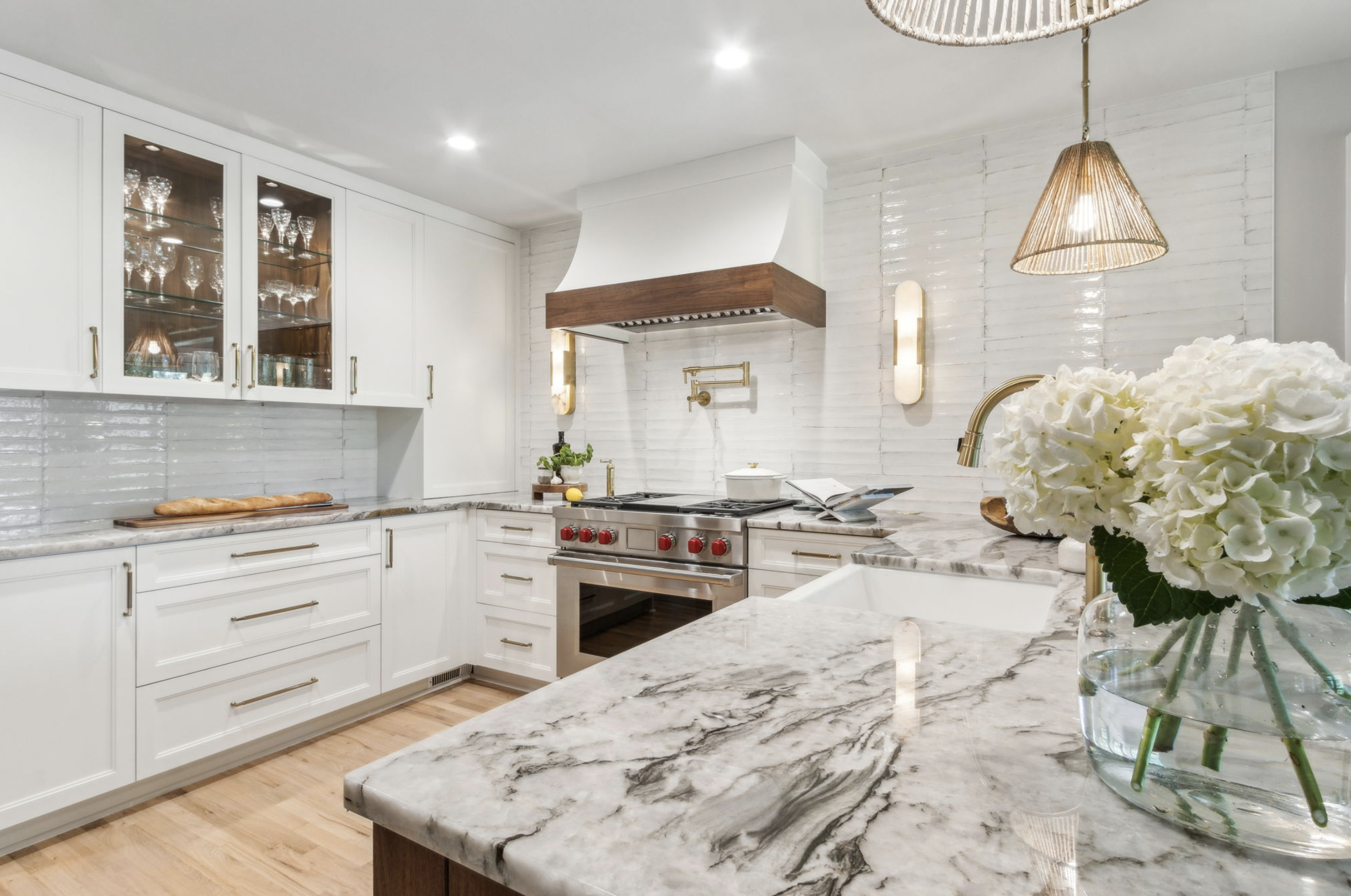 Modern kitchen with white cabinets, marble countertops, stainless steel oven, and decorative lighting, with a vase of white hydrangeas on the island.