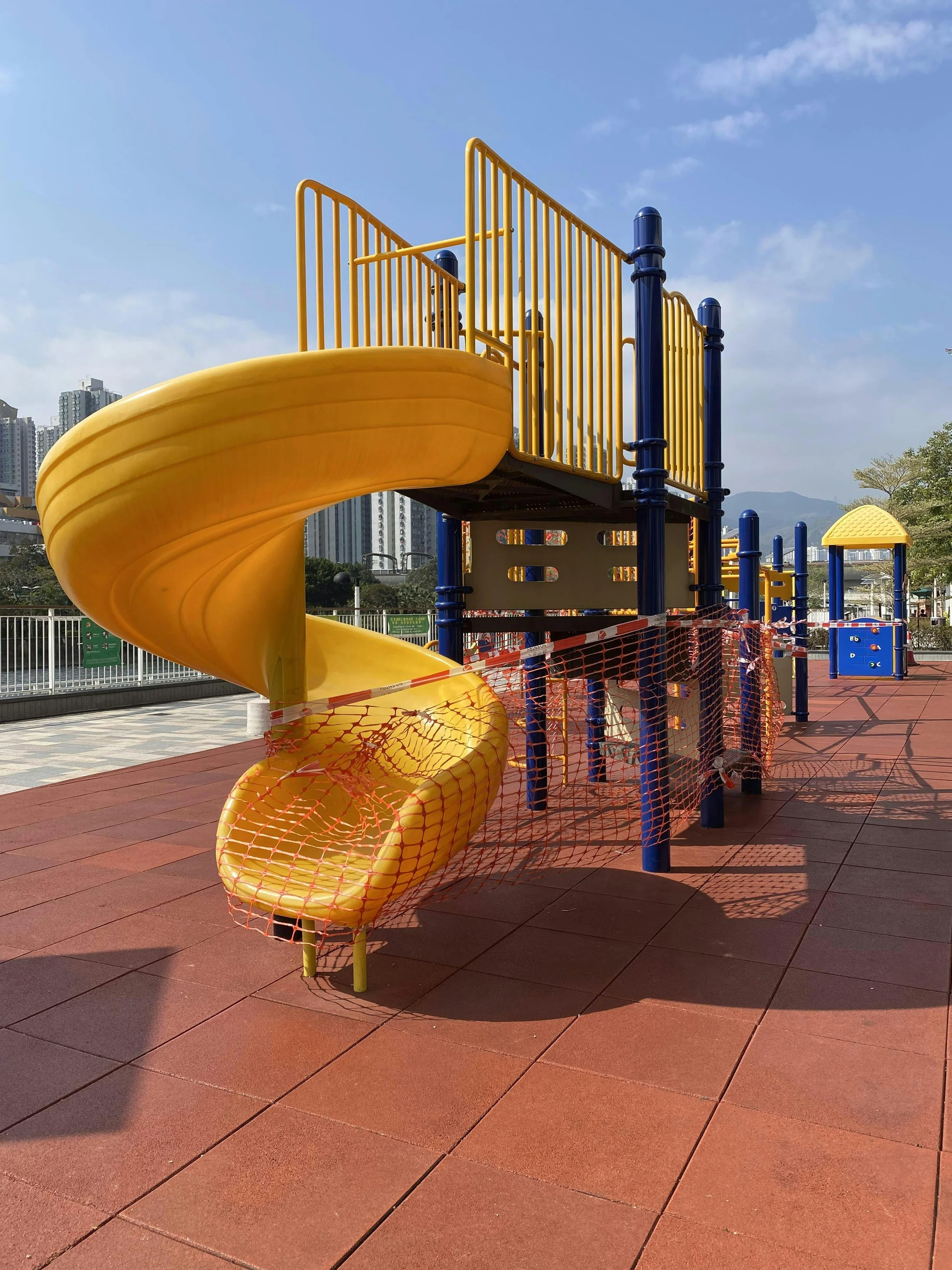 Children's yellow spiral slide on colorful playground with blue and yellow play structures and safety netting, outdoor setting with cityscape and mountains in background