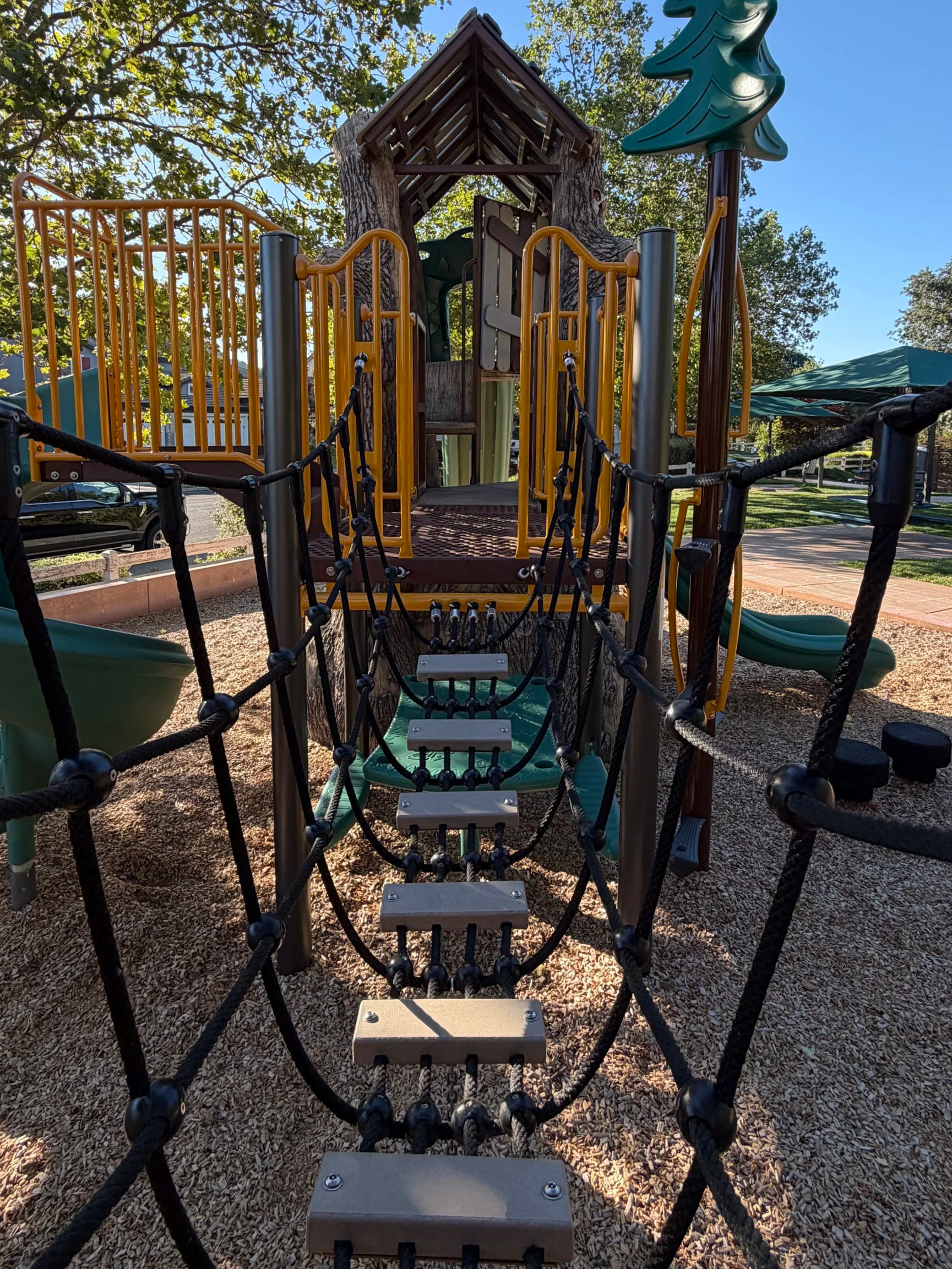 A playground designed as a treehouse with a rope bridge and slides, set in a park with trees and clear blue sky.