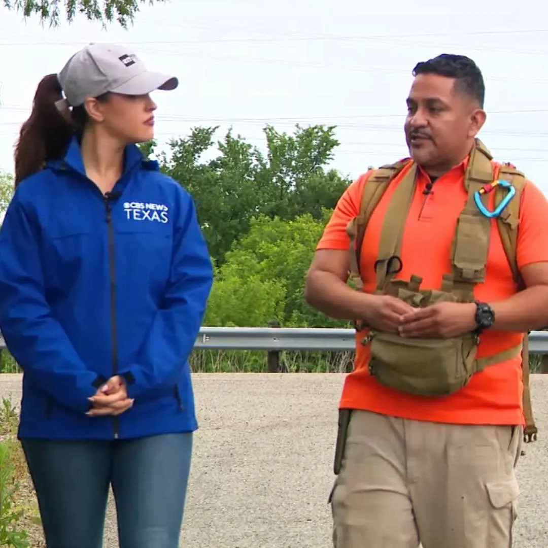 A female news reporter in a blue jacket and gray cap speaking with a male hiker in an orange shirt and hiking gear outdoors with greenery in the background.