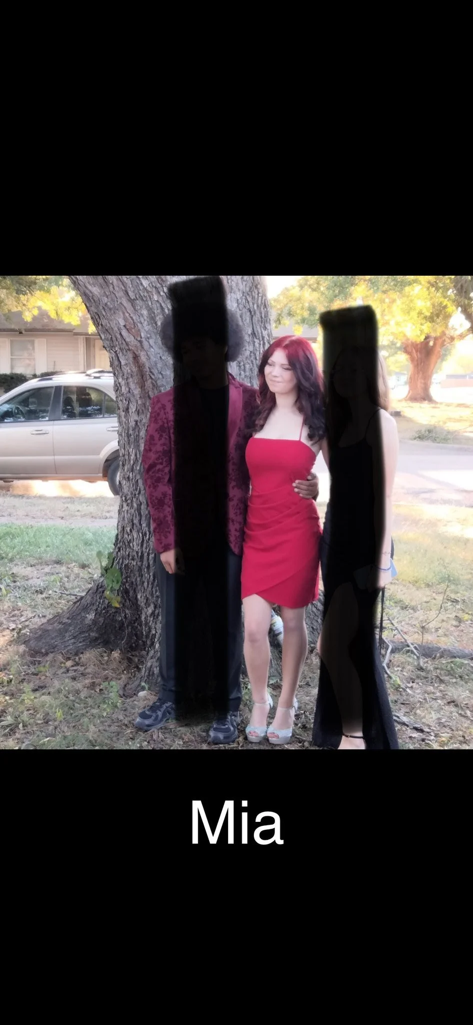 A photo of three people standing outdoors on grass in front of a large tree, with a car and houses visible in the background. The person in the middle is a woman wearing a red dress and high heels, smiling with her eyes closed. To her left is a man w