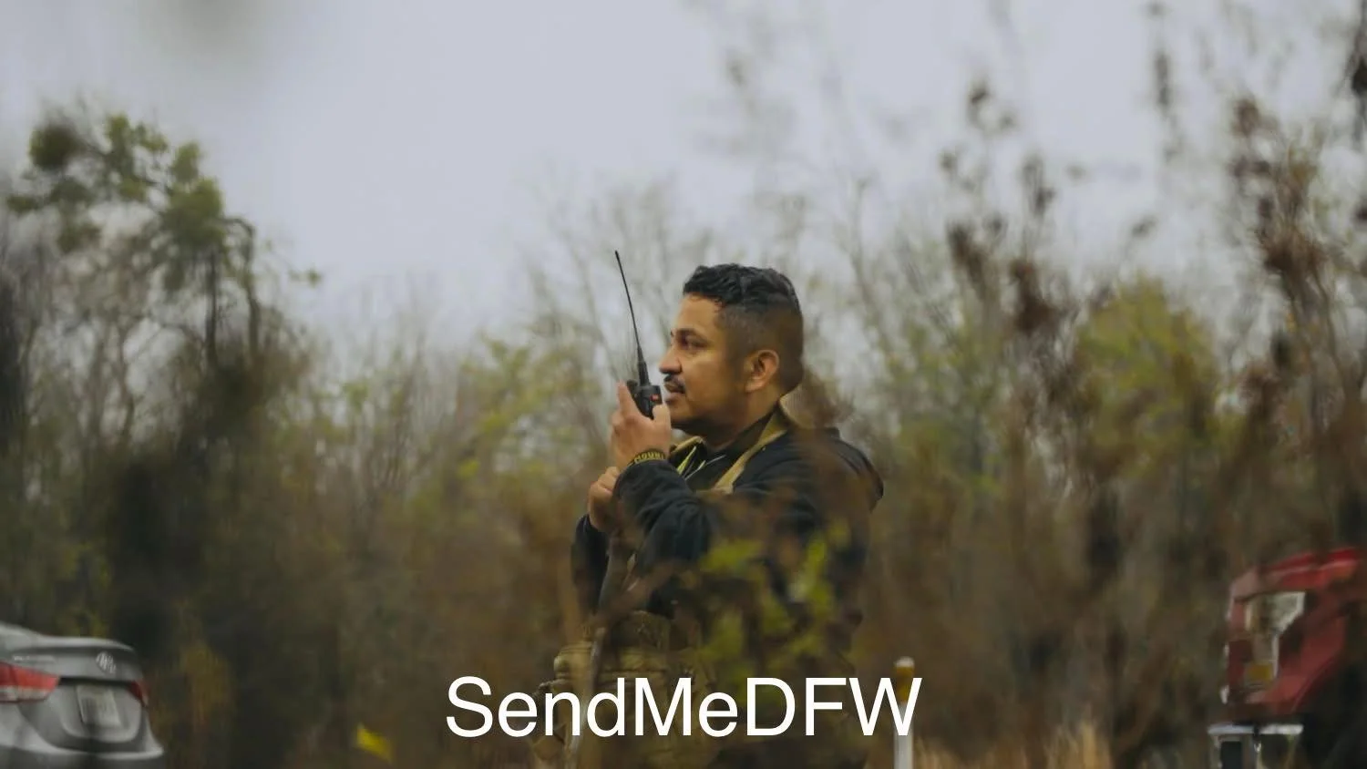 A man standing outdoors in a rural area, holding a walkie-talkie near his face while looking to the left. The background features blurred trees and bushes with an overcast sky.