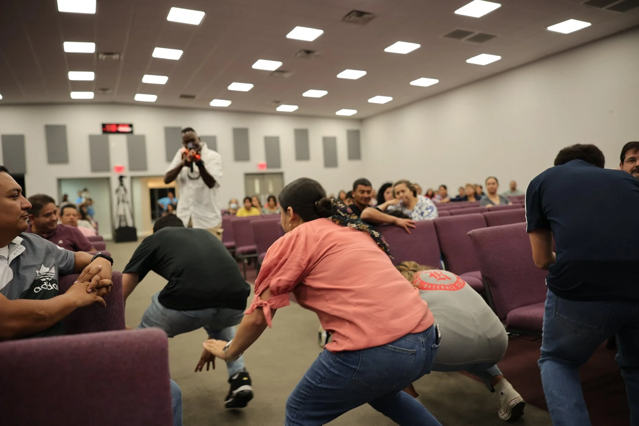 People seated in a church or auditorium with some standing and others sitting, as a woman leans forward and reaches out during a gathering, with a man in a white coat taking a photo in the background.