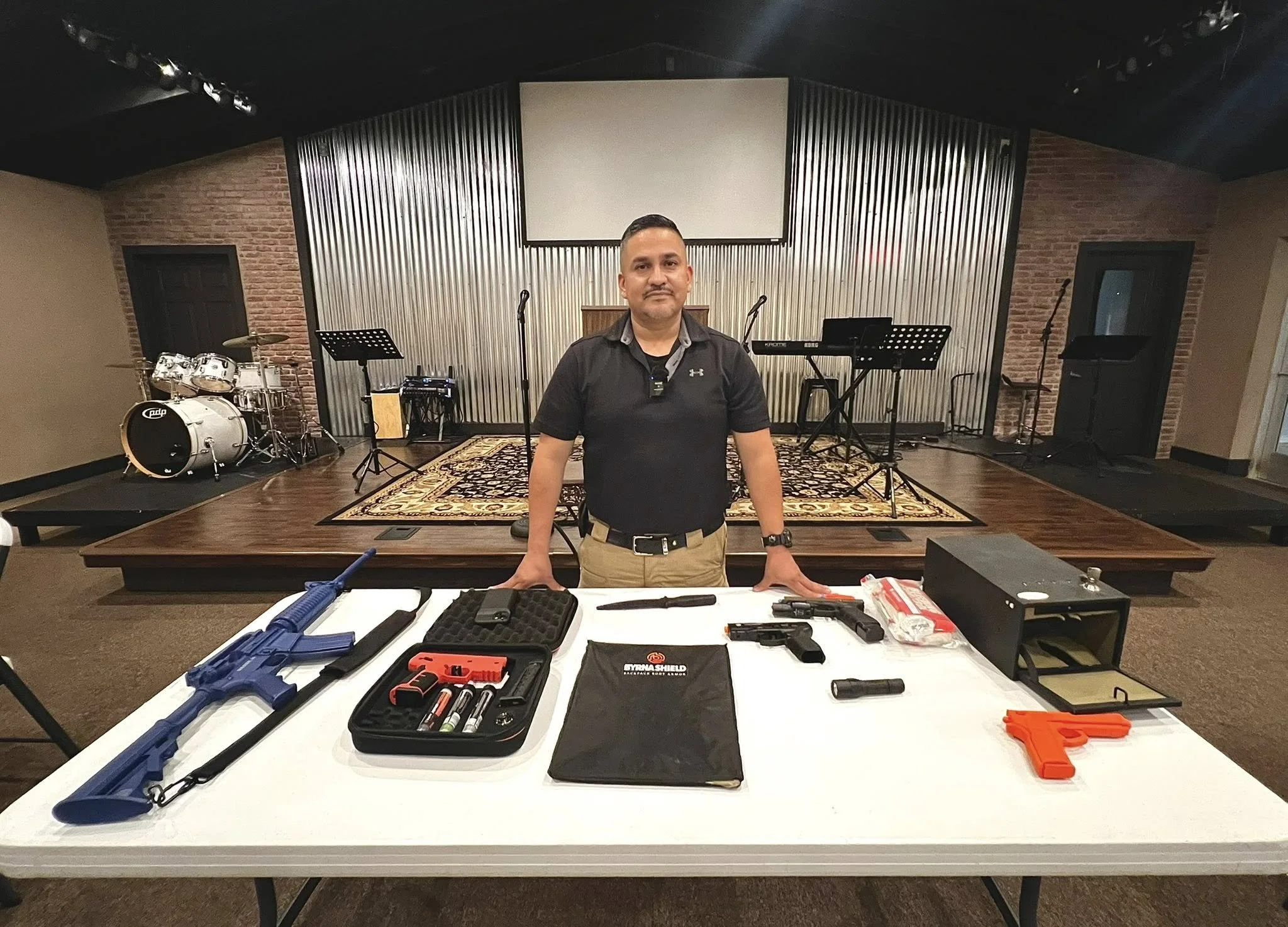 Man standing behind a table with firearms and ammunition, in a room with musical instruments on stage behind him.