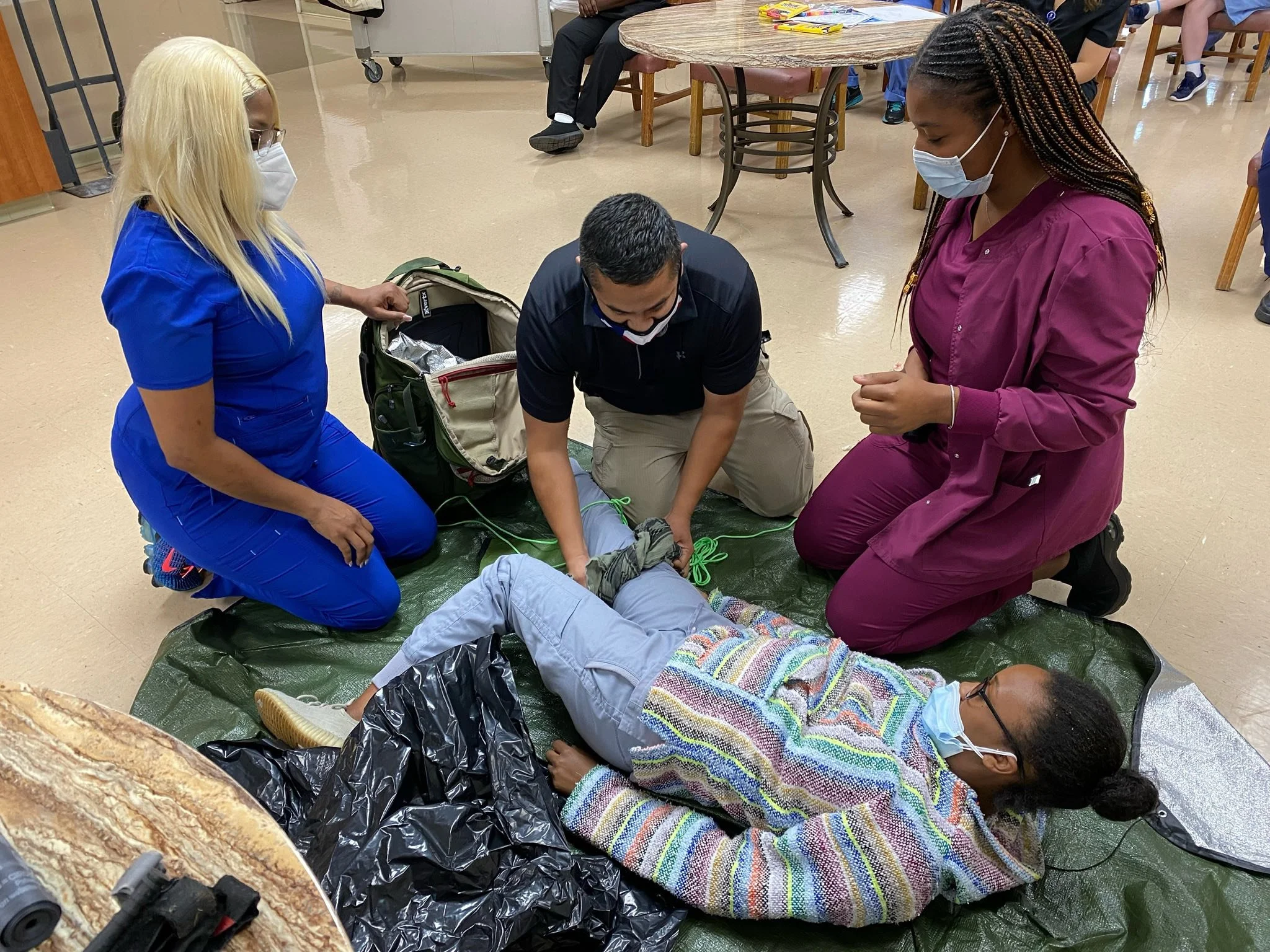 Three healthcare professionals attending to a person lying on a mat, practicing emergency medical procedures. All wear masks and gloves, and the setting appears to be a training session or medical facility.