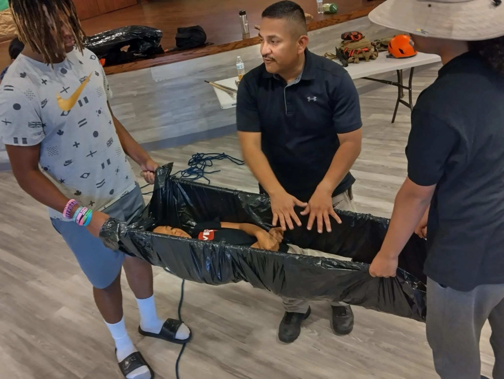 Three people are holding a stretcher with a person lying down inside, in an indoor setting with tables and safety gear in the background.