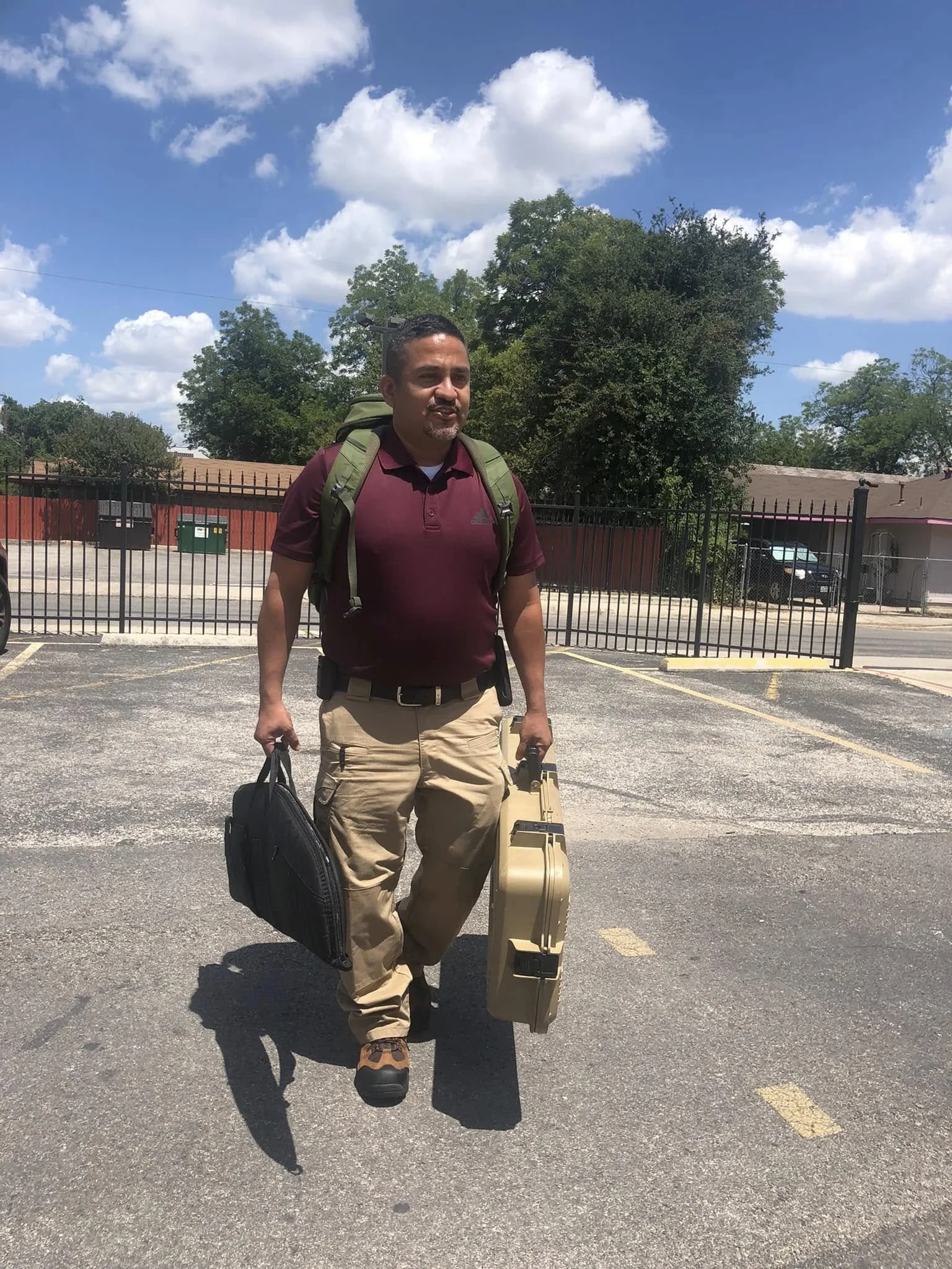 A man walking through a parking lot with a green backpack, carrying a black bag in one hand and a tan briefcase in the other. He is wearing tan cargo pants, a maroon polo shirt, and brown shoes. The background shows a black fence, some trees, and a partly cloudy sky.