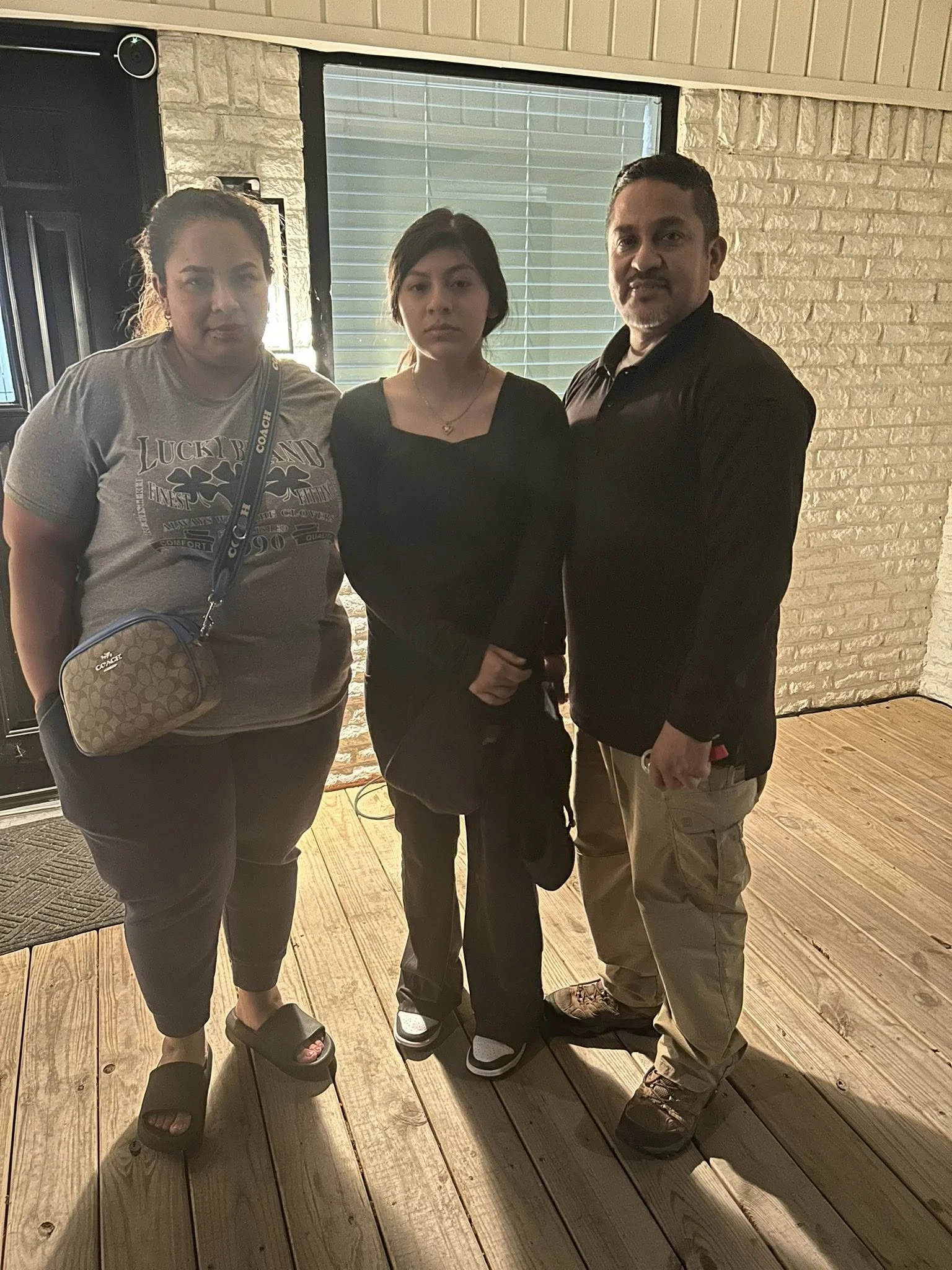 Three people standing indoors near a window, with a brick wall in the background and a wooden floor.