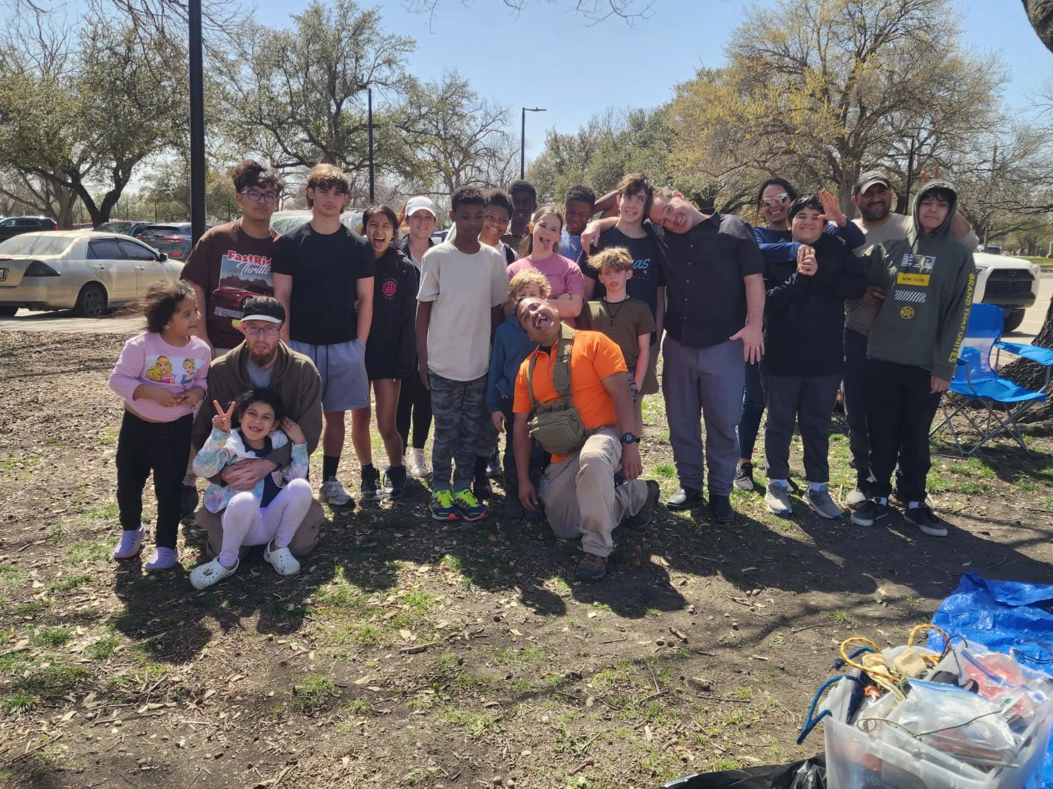 Group of diverse children and adults outdoors on a sunny day, posing for a group photo during a community event, with some smiling and others making playful gestures.
