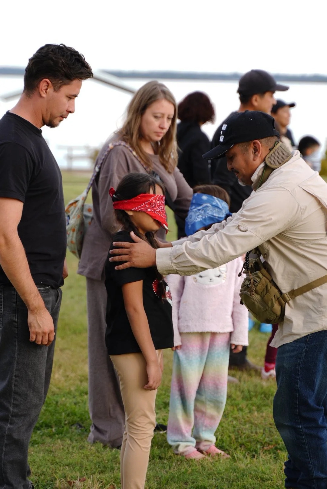 A group of people, including children with bandanas over their eyes, participate in a trust exercise outdoors, led by a man.