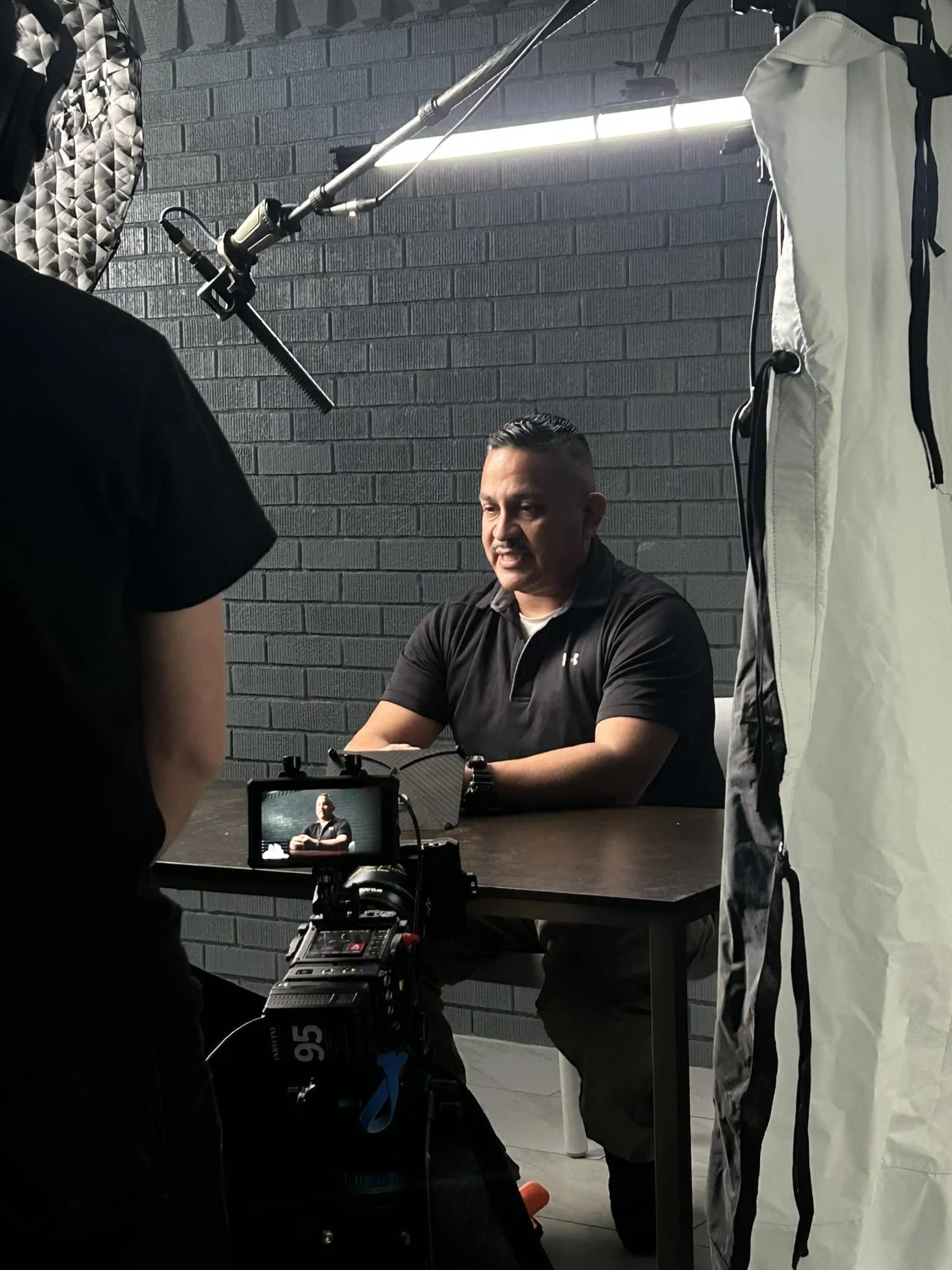A man sitting at a table during a video shoot or interview, with film equipment, camera, and lighting setup in a room with dark brick wall background.
