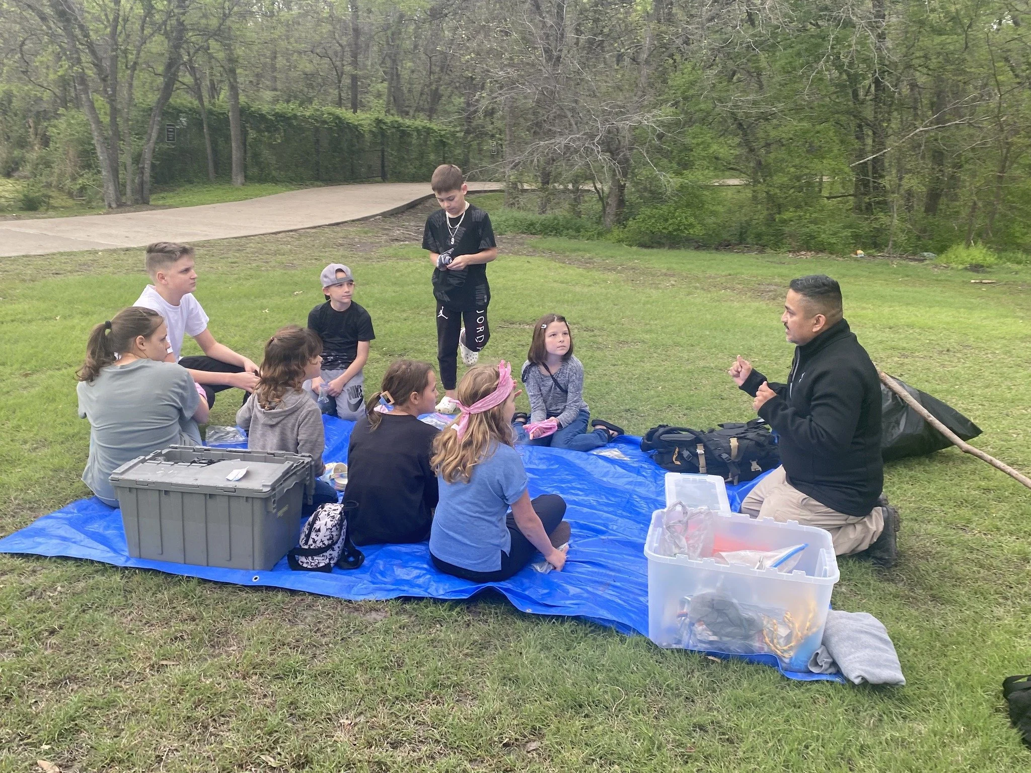 A group of children sitting on a blue tarp listening to a man in a black jacket speaking outdoors in a park.