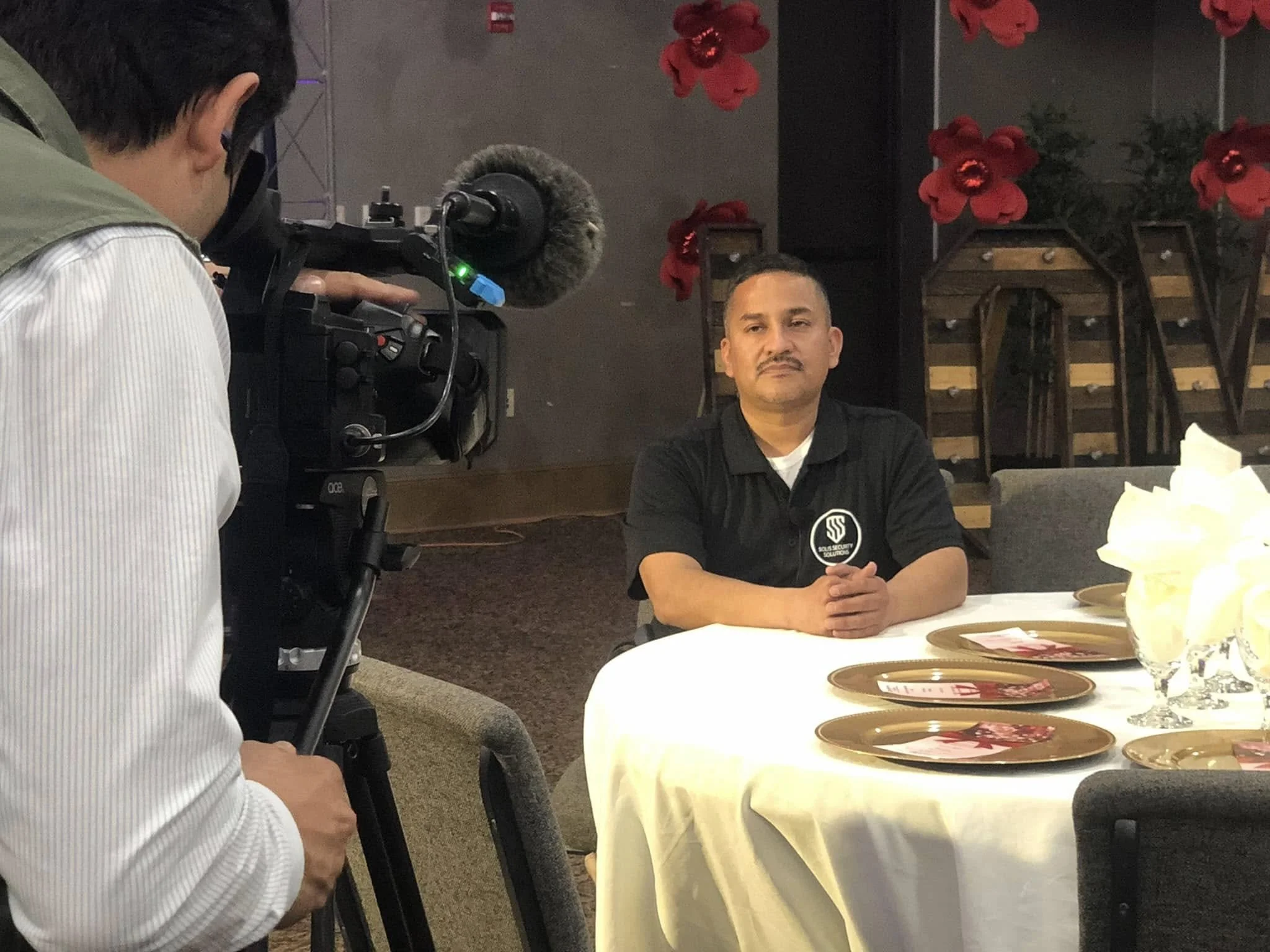 A man is sitting at a round table during a video recording or interview. A cameraman with a professional camera is filming him. The man is wearing a black polo shirt with a logo, and he has his hands rested on the table. The table is set with golden 