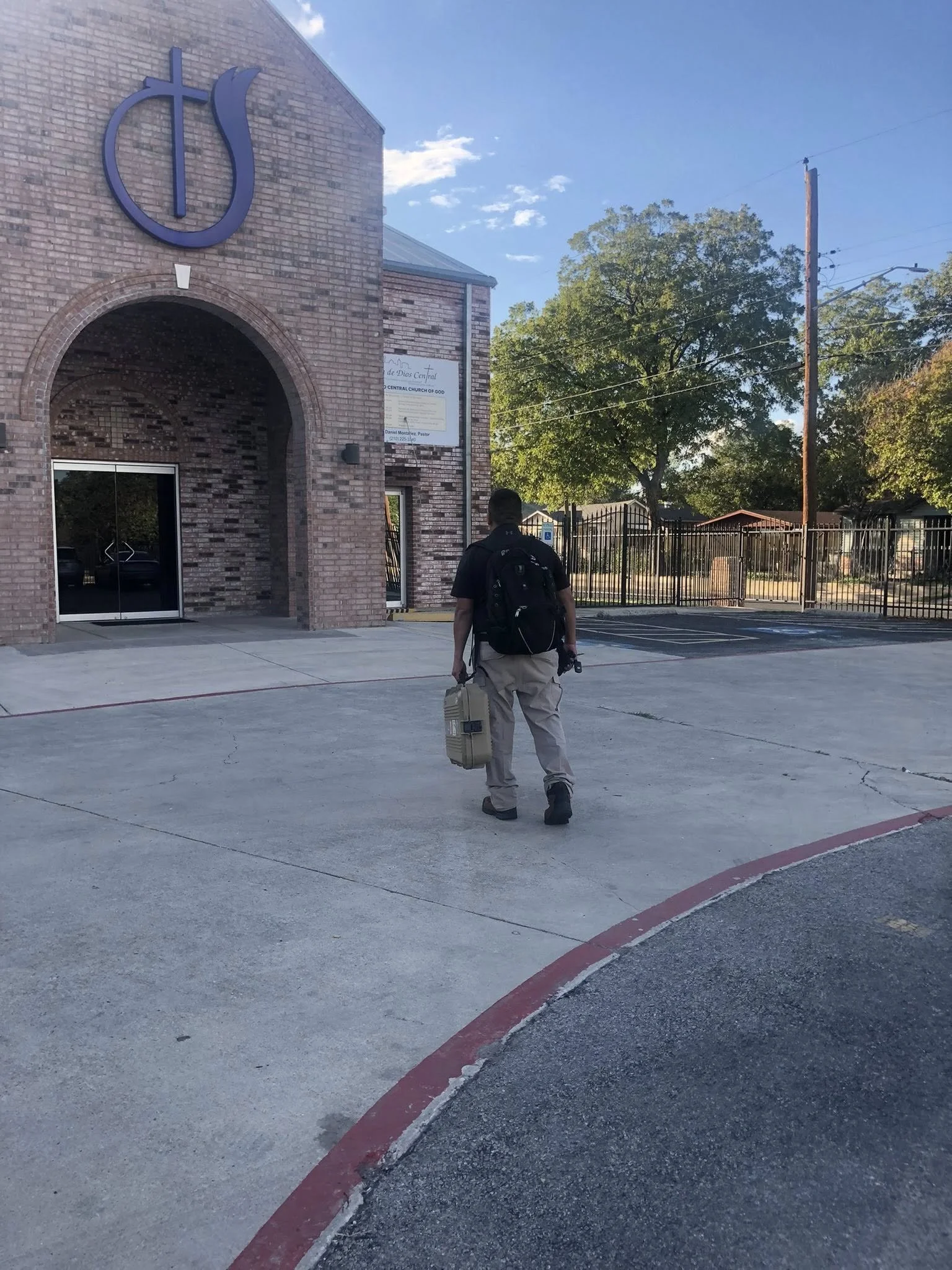 A man with a black backpack and beige pants walking towards the entrance of a brick church, carrying a beige case in his right hand. The church has a large blue symbol above the arched doorway, and a small sign on the side. There are trees and a meta