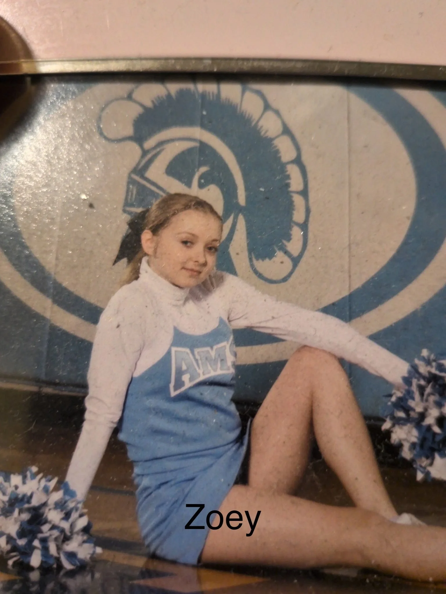 A young woman in a cheerleader uniform sitting on a gym floor with a pom-pom in her hand, in front of a wall with a large blue Spartan helmet logo. The name 'Zoey' is written at the bottom of the image.