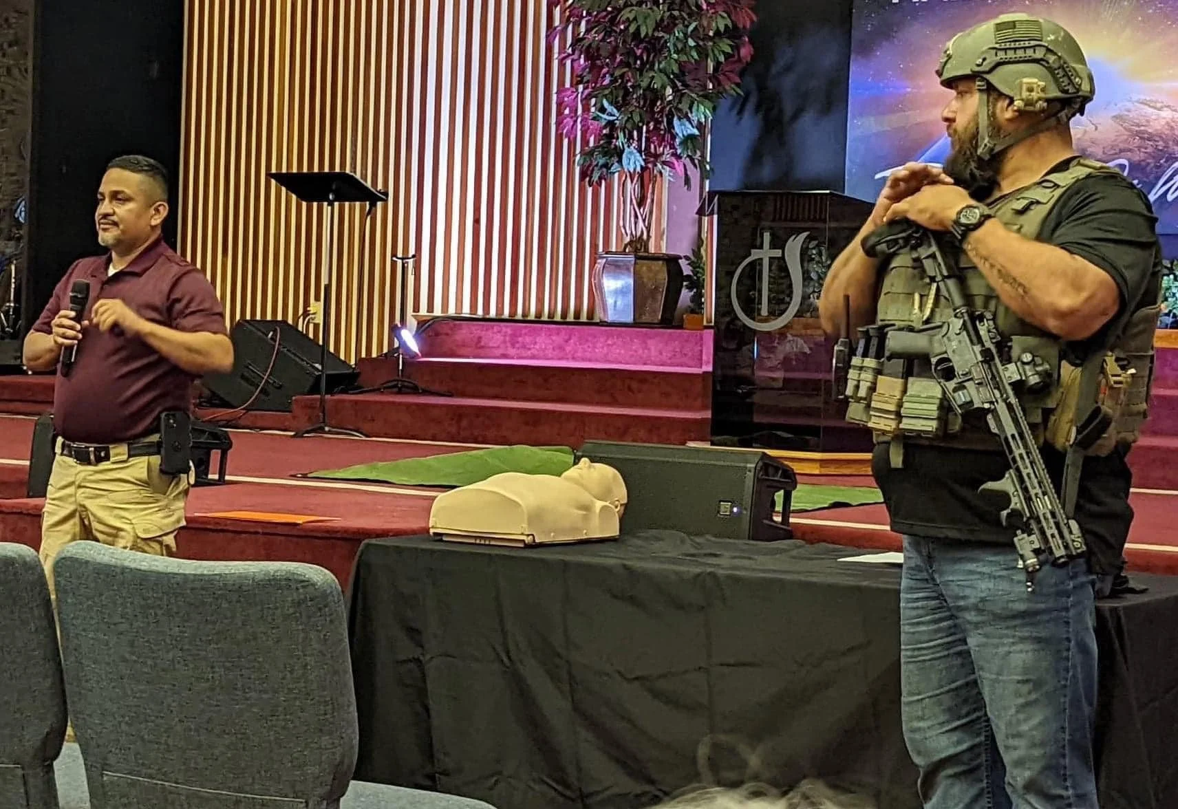 A man in a red shirt holding a microphone and a man in tactical gear with a rifle are engaged in a presentation in a church. There is a CPR mannequin on a table in front of them, with a church stage in the background.