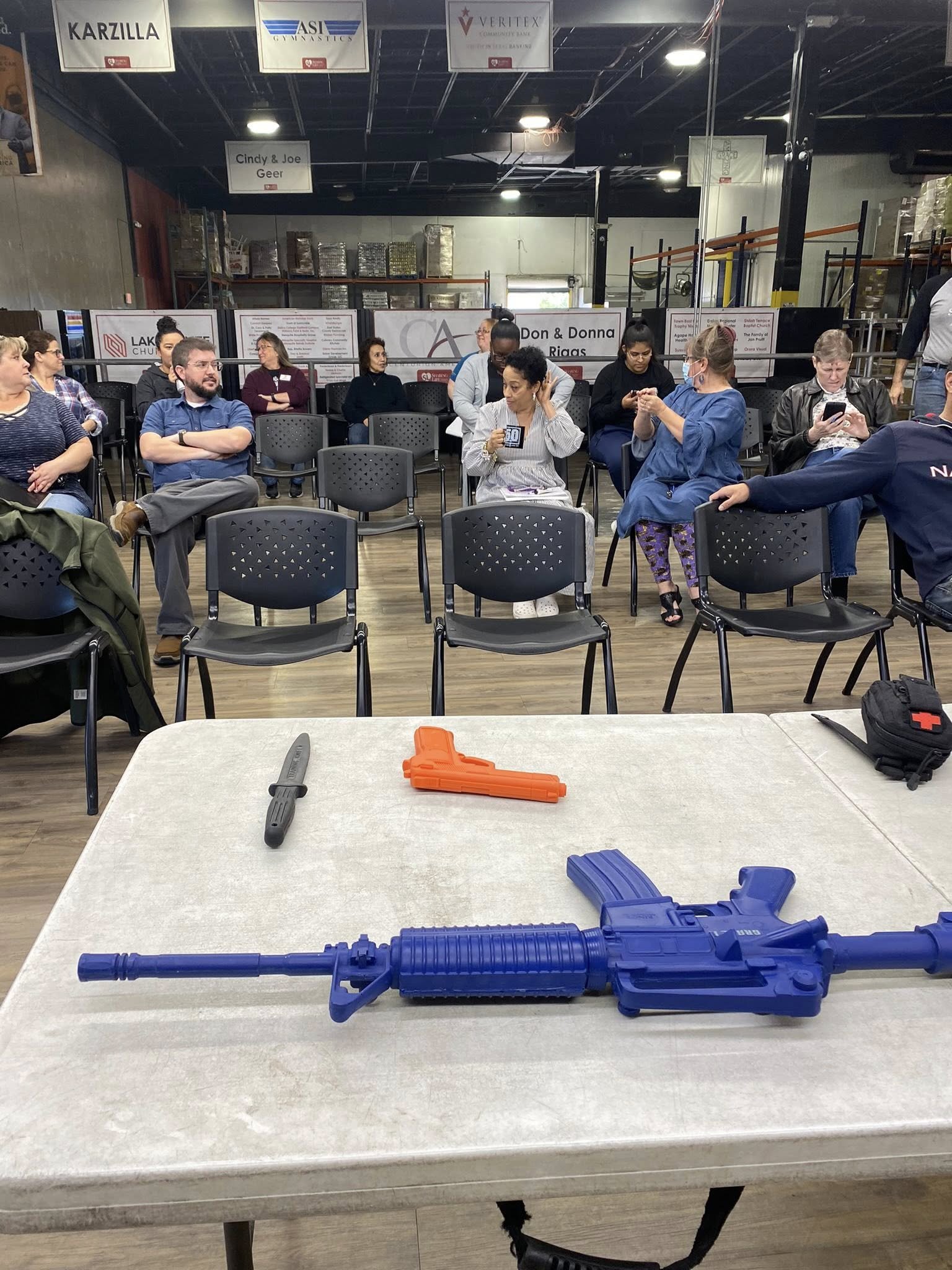Table with toy gun and knife in the foreground, people sitting and waiting in chairs in a community center or gymnasium in the background.