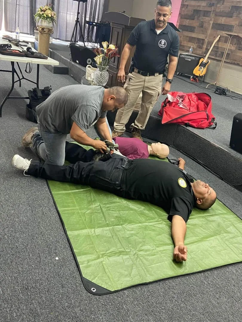 Two men practicing CPR on a training mannequin lying on a green mat while another man observes in a room with musical instruments and flowers.