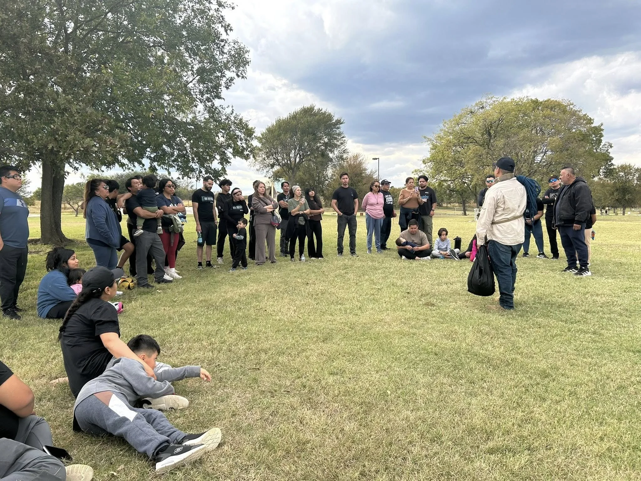 Group of people gathered outdoors in a park, listening to a man speaking with a bag, surrounded by trees and a cloudy sky.