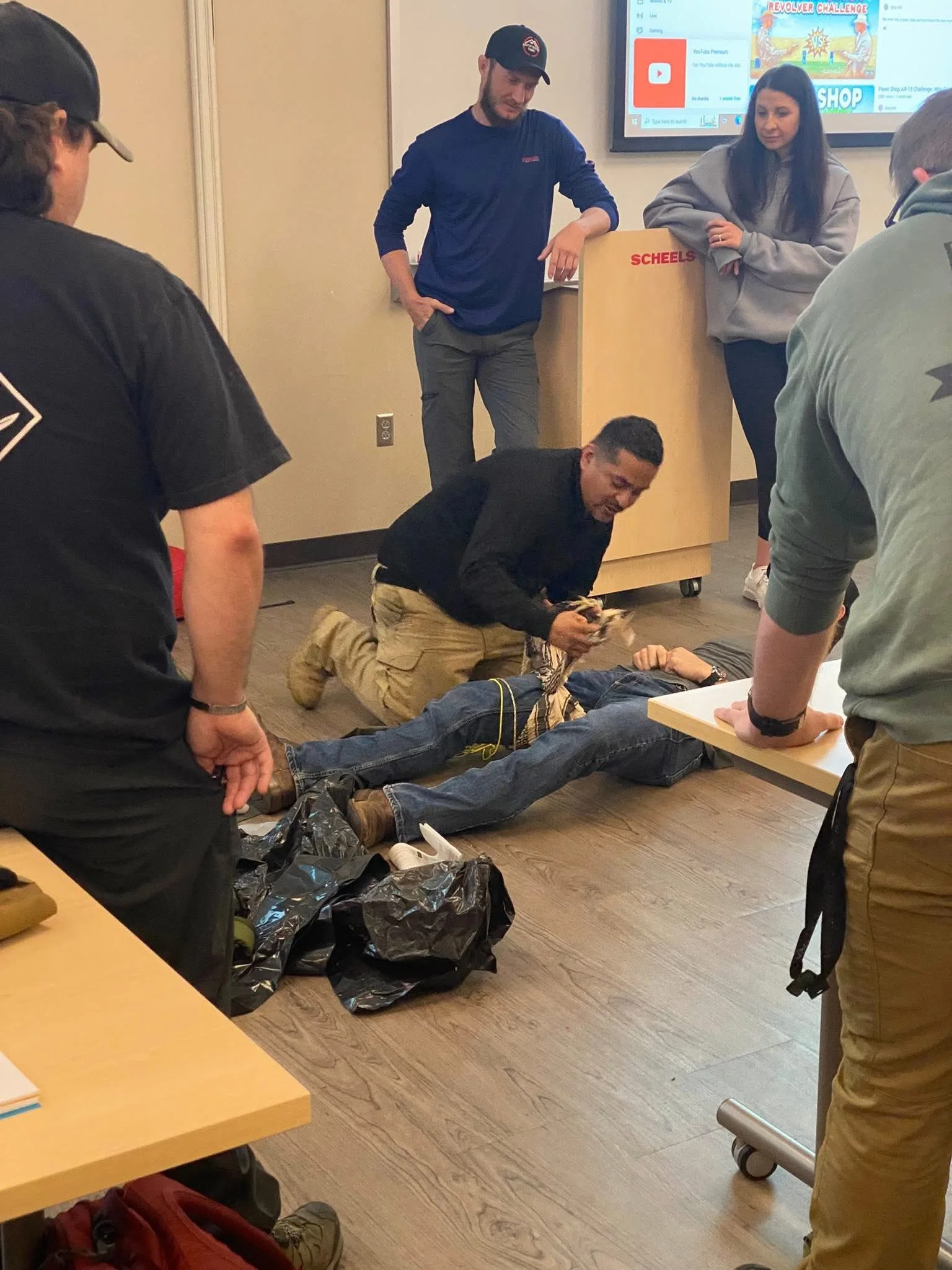 Group of people practicing first aid or emergency response training on a person lying on the floor. One person is kneeling and holding a cloth or bandage, while others are watching and observing. The setting appears to be a classroom or training room with a large screen displaying a YouTube page in the background.