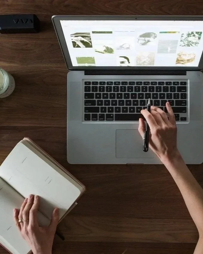 A person working on a laptop at a wooden desk with an open notebook and a small plant on the side.