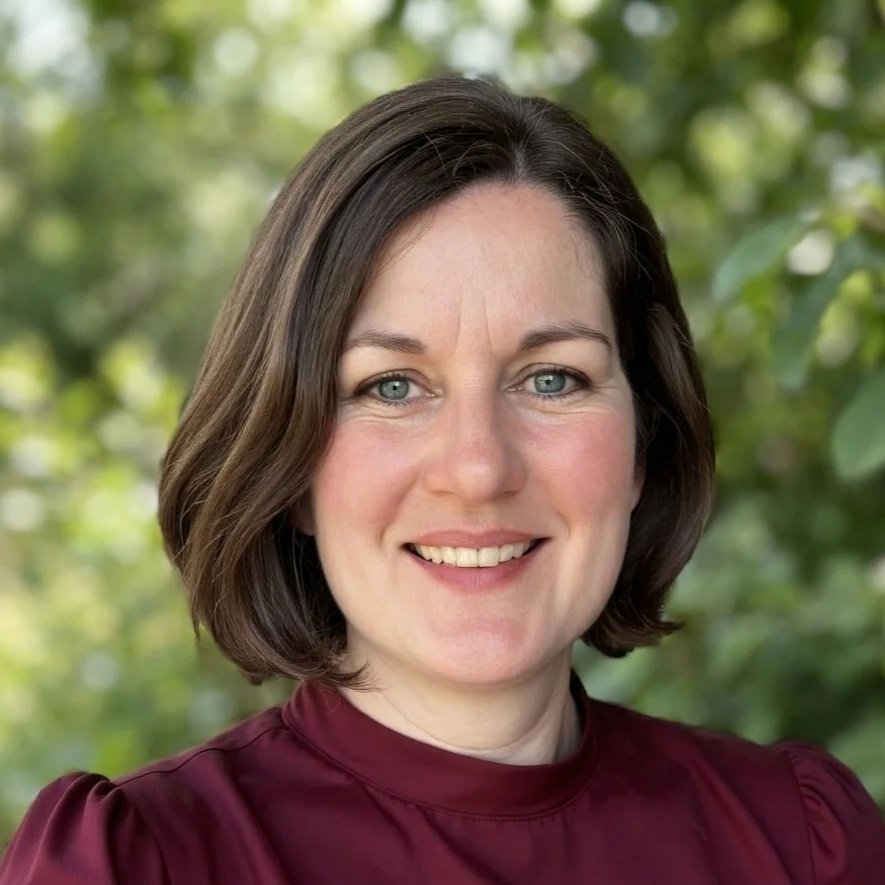 Portrait of a woman with short brown hair, blue eyes, smiling, wearing a burgundy top, outdoors with green foliage in the background.