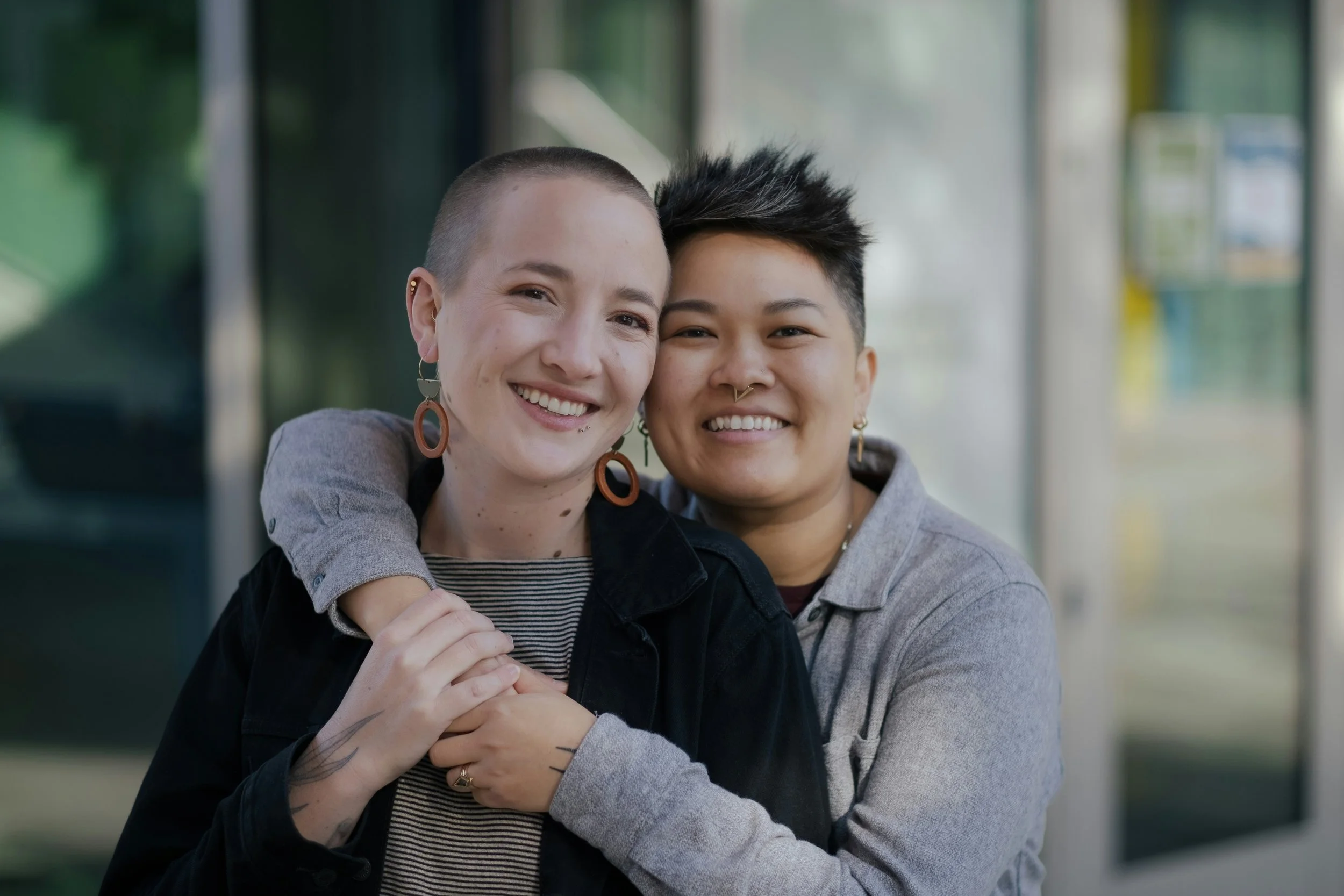 Two smiling women hugging each other outdoors, one with short dark spiky hair and a nose piercing, the other with very short shaved hair, wearing earrings and a striped shirt underneath a dark jacket.