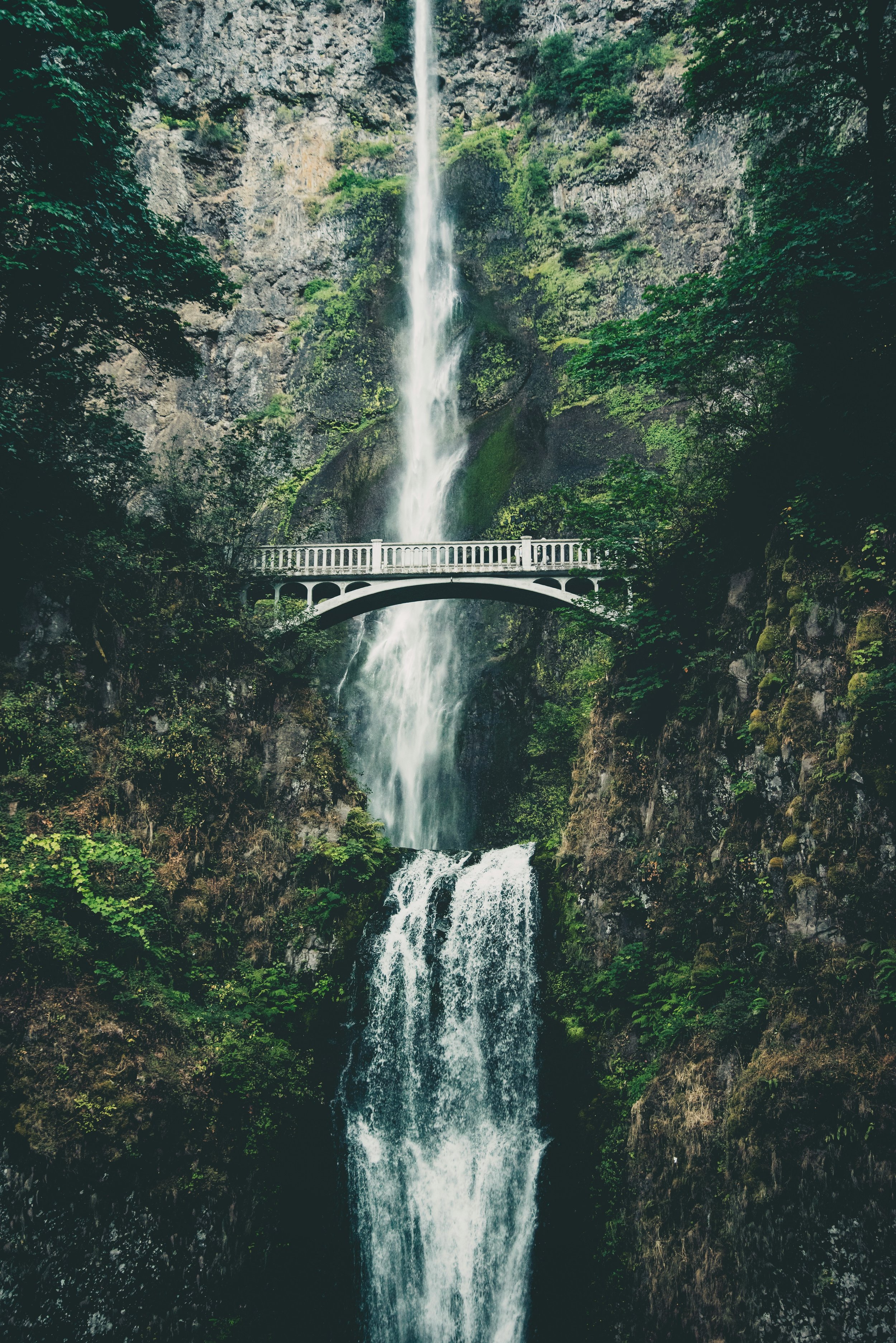 A tall, narrow waterfall cascading down a rocky cliff surrounded by lush green trees, with a white bridge across the waterfall.