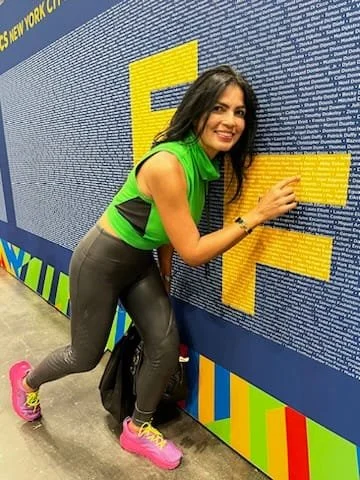 Woman with long dark hair wearing a green and black athletic shirt, black leggings, and pink sneakers, smiling while pointing at a large text wall at an event.