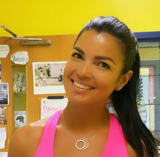 Smiling woman with dark hair in a ponytail, wearing a pink top and silver necklace, indoors.