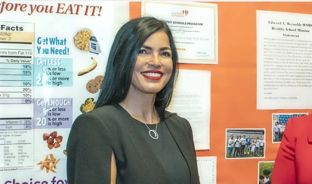 A woman with long dark hair and red lipstick standing in front of an orange bulletin board with educational posters and photos.