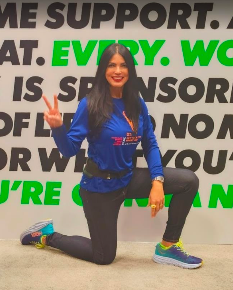 A woman smiling and posing in front of a wall with motivational text, making a peace sign with her right hand, wearing a blue athletic jacket, black pants, and running shoes.
