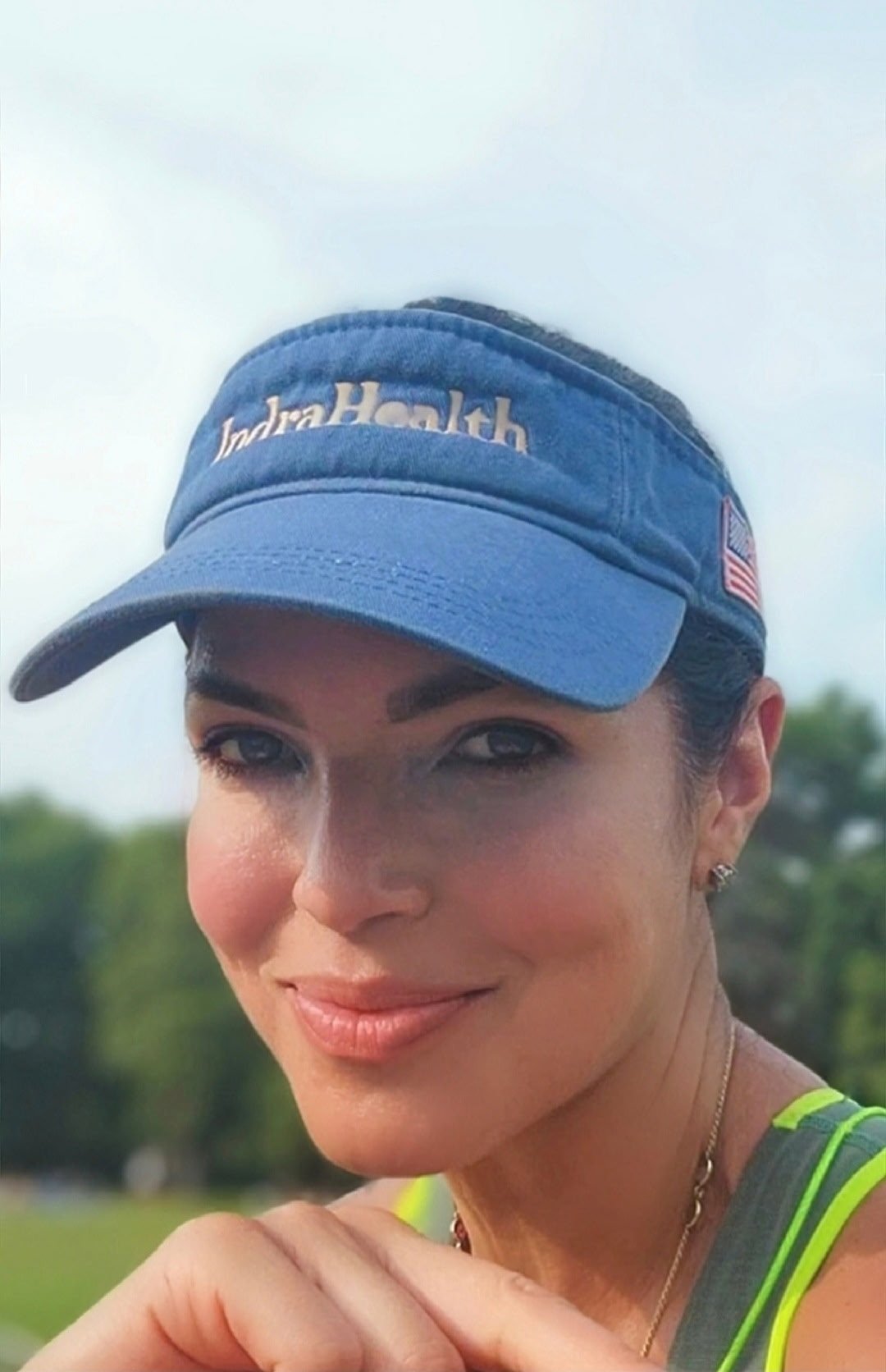A woman taking a selfie outdoors, wearing a black visor, red athletic top, and white earphones, with a road and green landscape in the background.