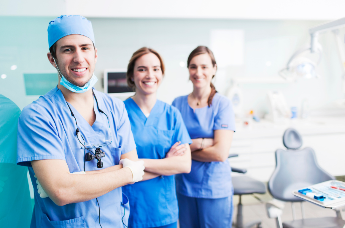 Three healthcare professionals in scrubs and caps standing in a medical clinic, smiling at the camera.