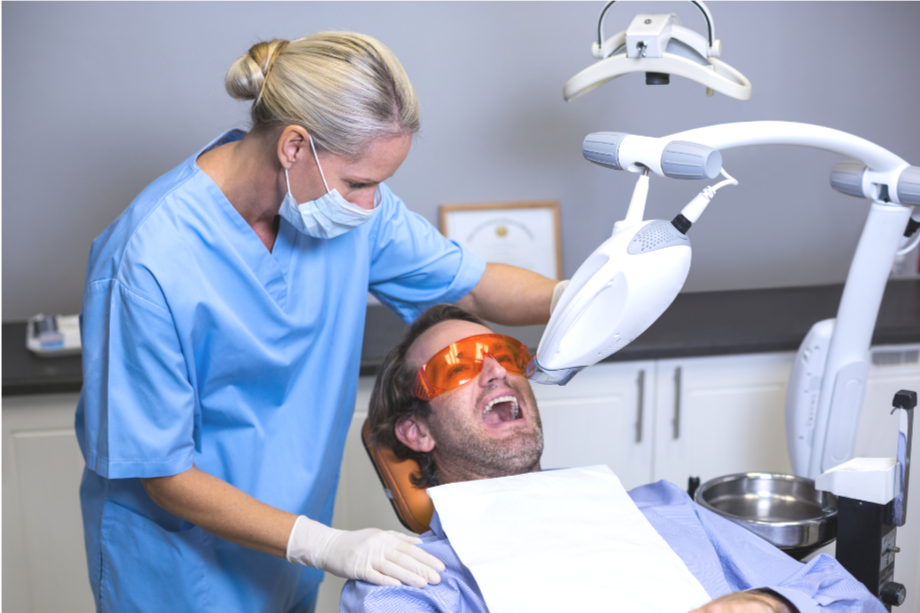 A dental professional wearing a face mask and gloves performing a procedure on a male patient lying in a dental chair, with a dental light and equipment in the background.