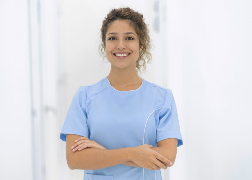 A smiling young woman in a light blue medical uniform standing with arms crossed in a bright, clean hospital corridor.