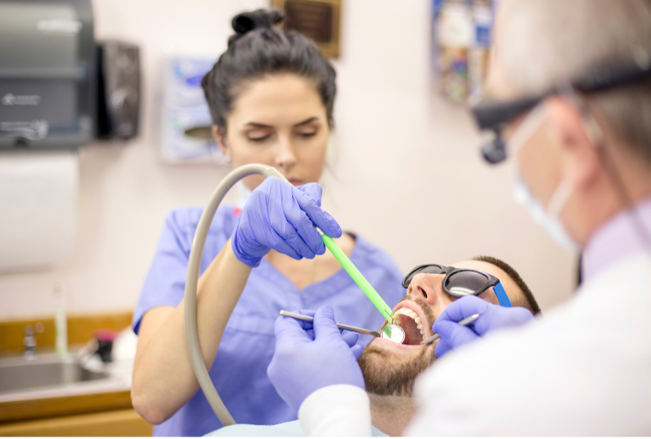 A dentist in protective glasses and gloves performs a dental checkup on a patient lying in the dental chair. The patient is wearing dark glasses and has his mouth open.