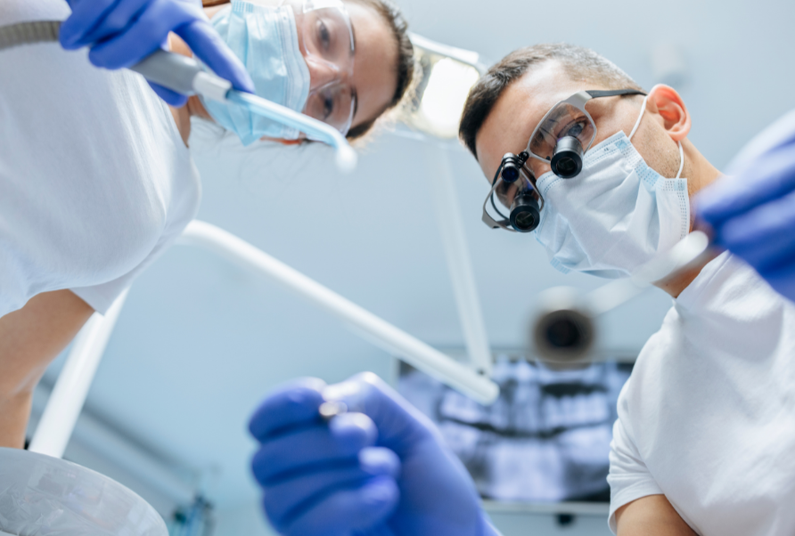 Two dentists wearing masks, gloves, and protective glasses work together on a patient in a dental office.