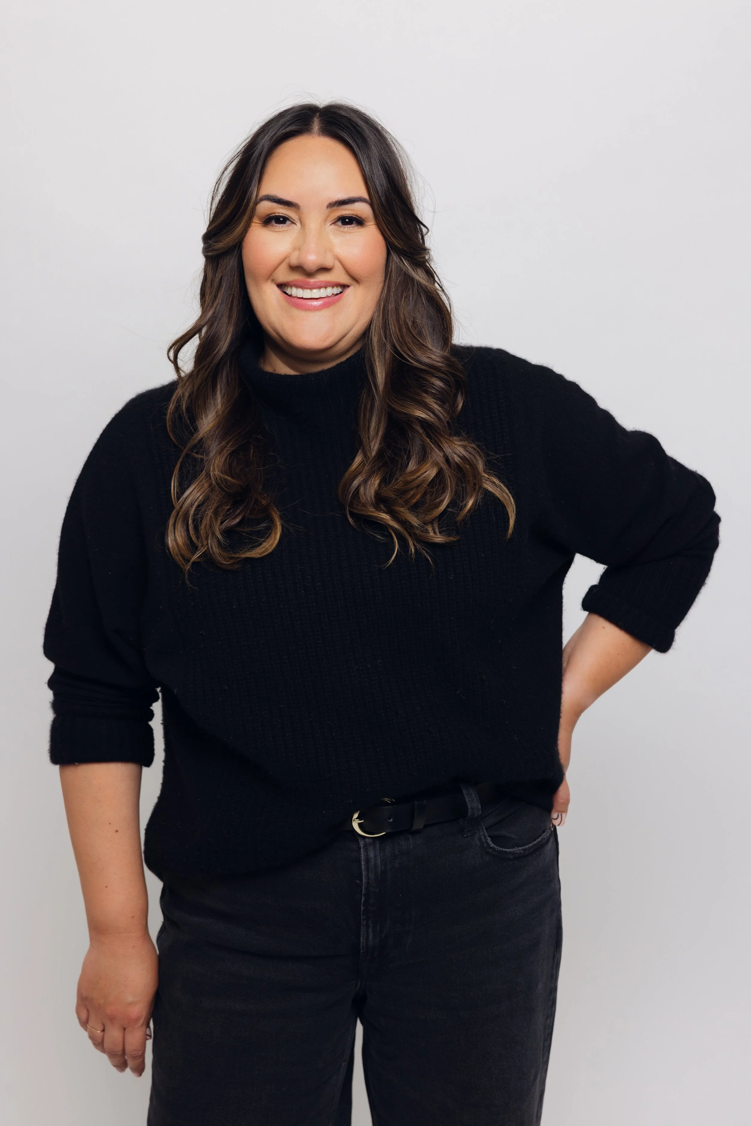 Woman with long, wavy brown hair and light skin, wearing a black sweater and black pants, smiling with her left hand on her hip, standing against a plain white background.