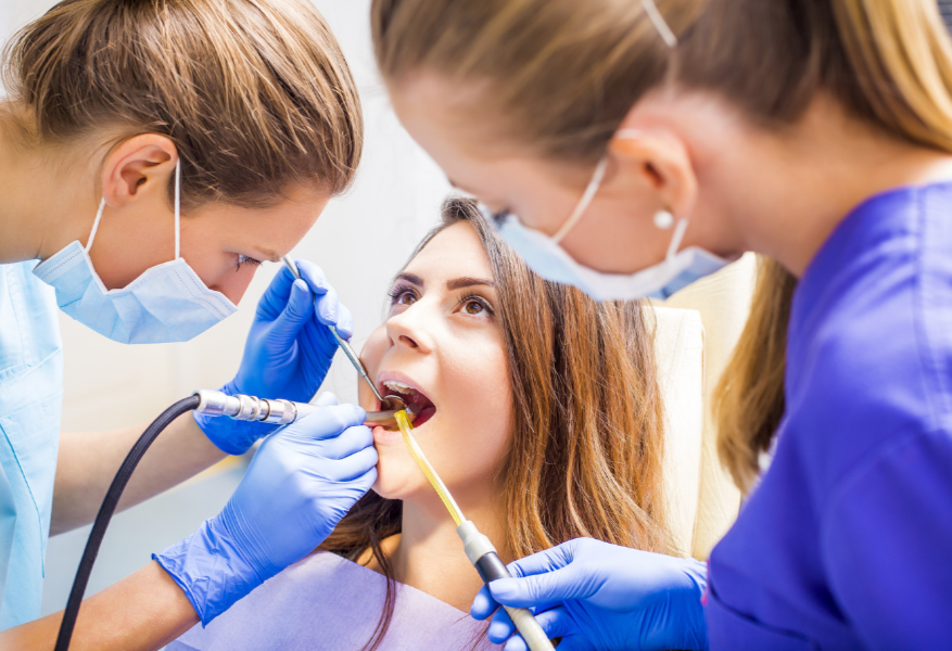 A woman receiving dental treatment from two dental professionals, both wearing masks and gloves in a dental clinic.