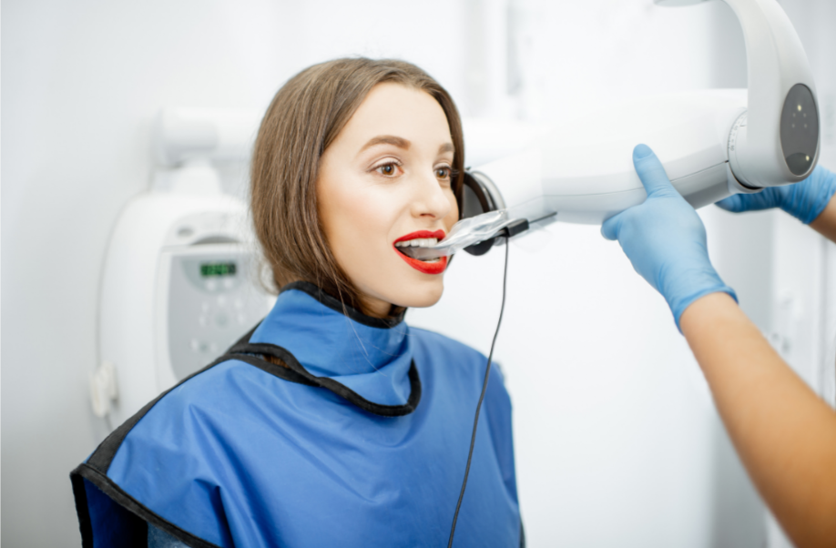 A woman is undergoing a dental X-ray scan with her mouth open, wearing a patient bib, while a technician in gloves positions the X-ray machine.
