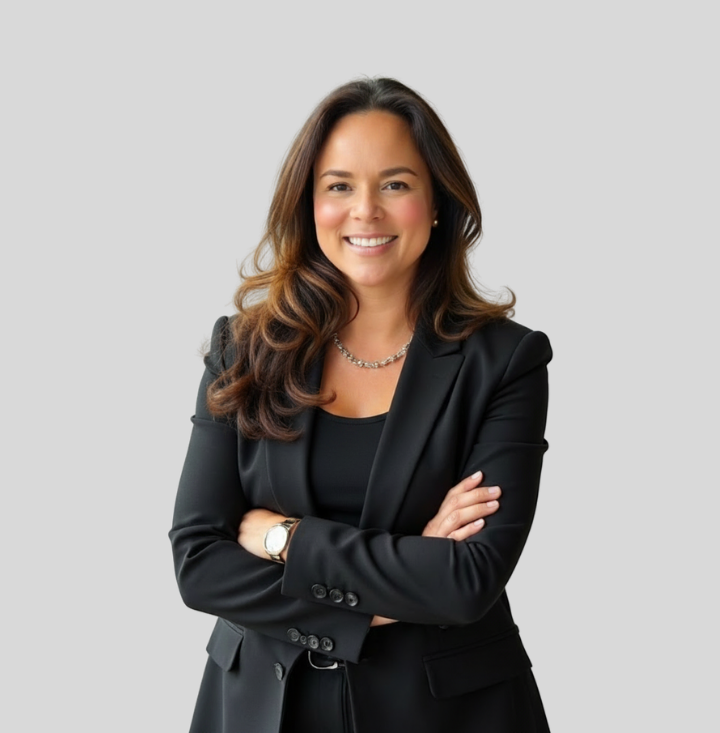A smiling woman with long brown hair wearing a black blazer, black top, and silver necklace, standing with crossed arms against a gray background.