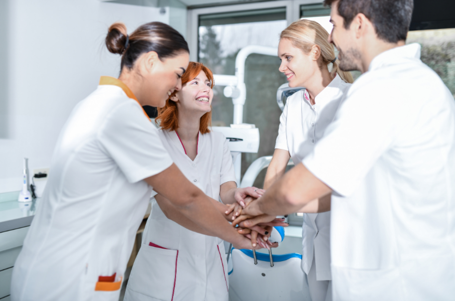 A group of five healthcare professionals in white medical uniforms holding hands together in a circle, smiling and celebrating in a modern clinic or hospital setting.